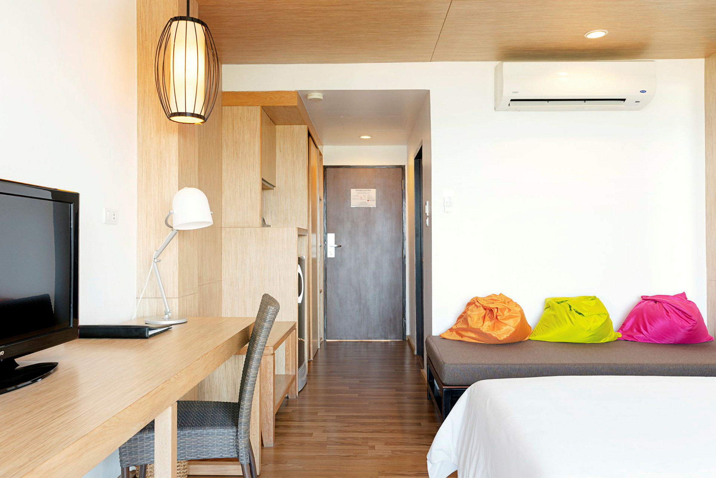 Ett hotellrum med skrivbord, stol, och TV. Färgglada sittsäckar är placerade på en soffa bredvid sängen. (AI-genererad text)
