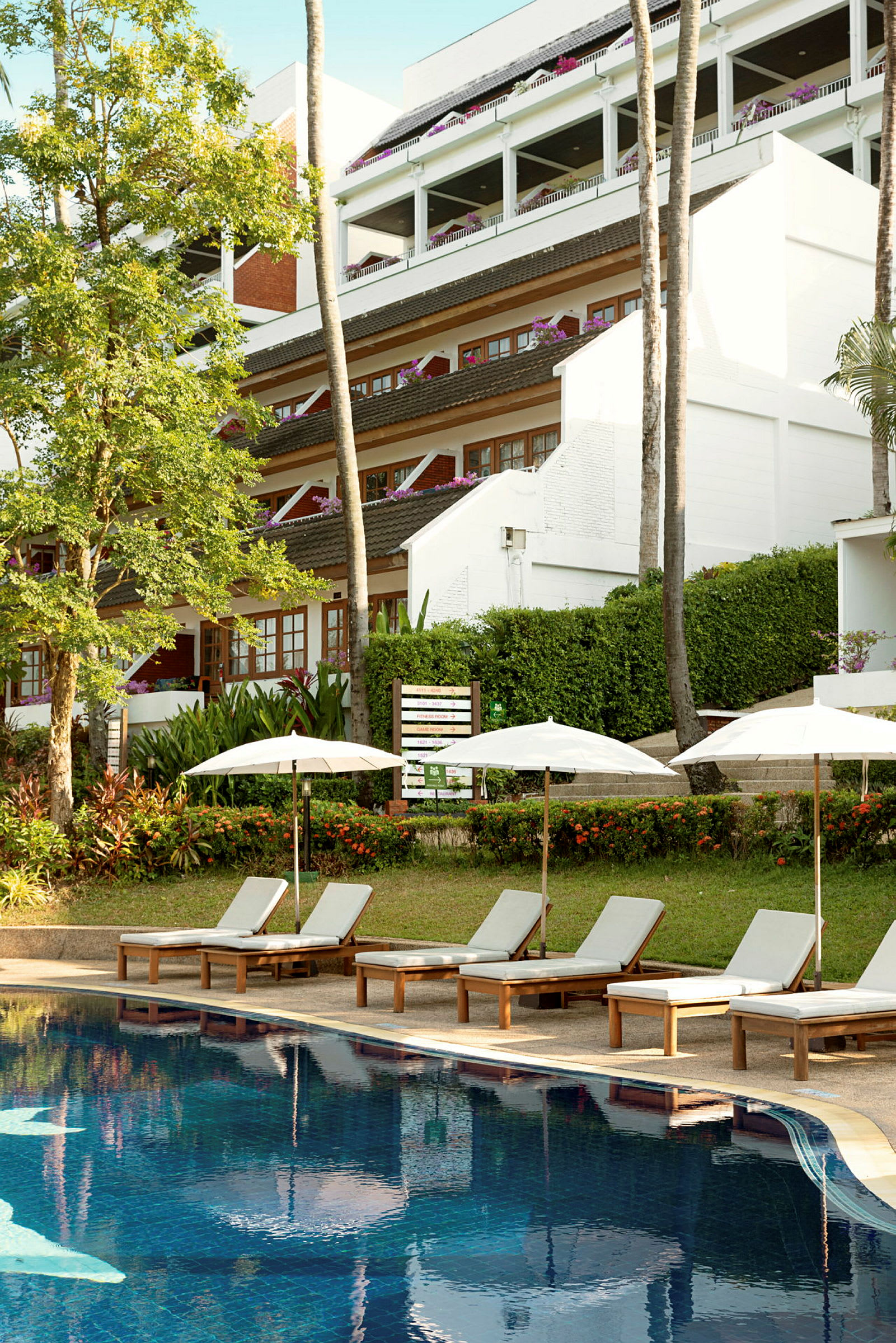 Solstolar och parasoller vid en pool, med grönska och hotellbyggnad i bakgrunden. (AI-genererad text)
