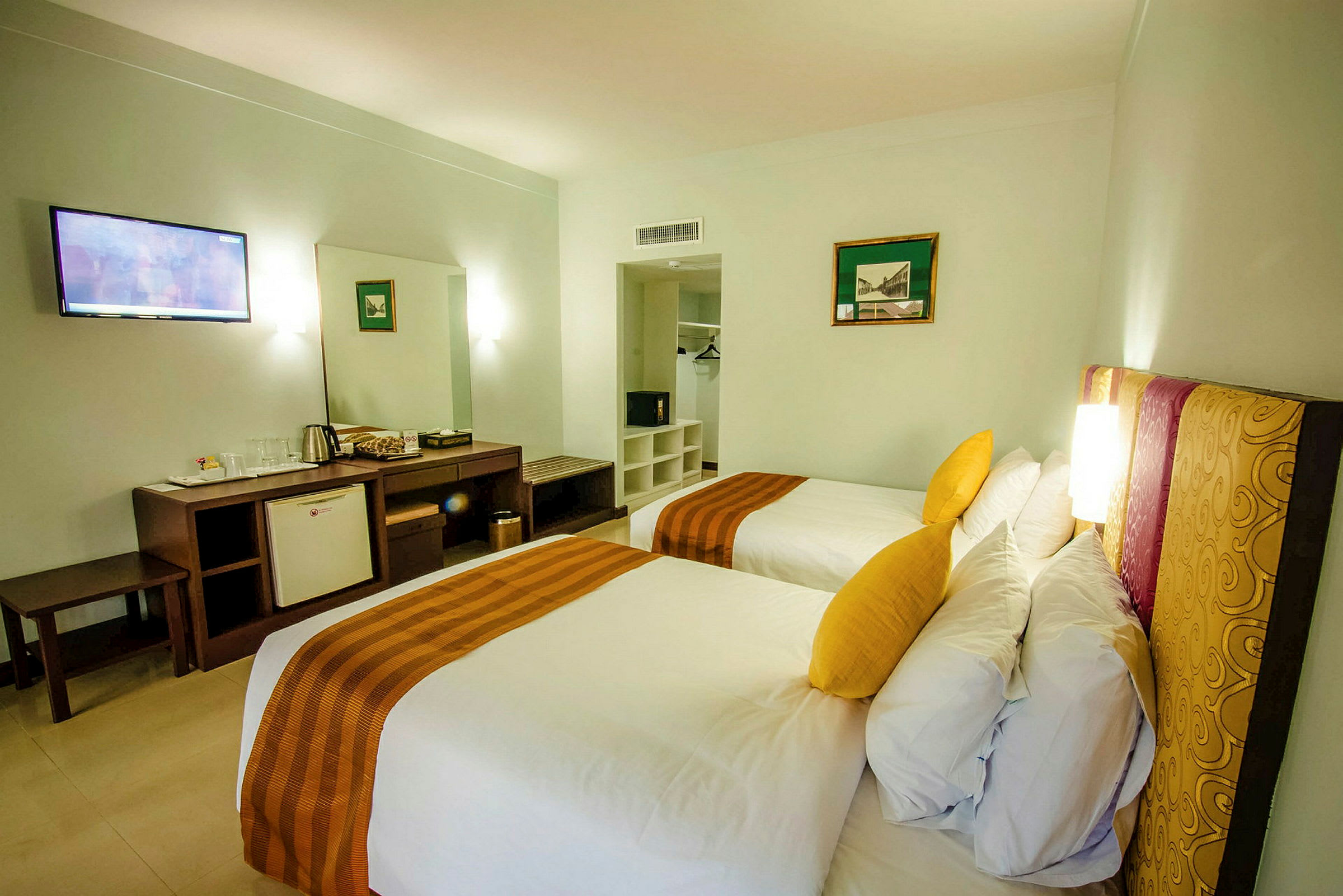 Två enkelsängar med gula kuddar i ett hotellrum, med skrivbord, minibar och garderob i bakgrunden. (AI-genererad text)
