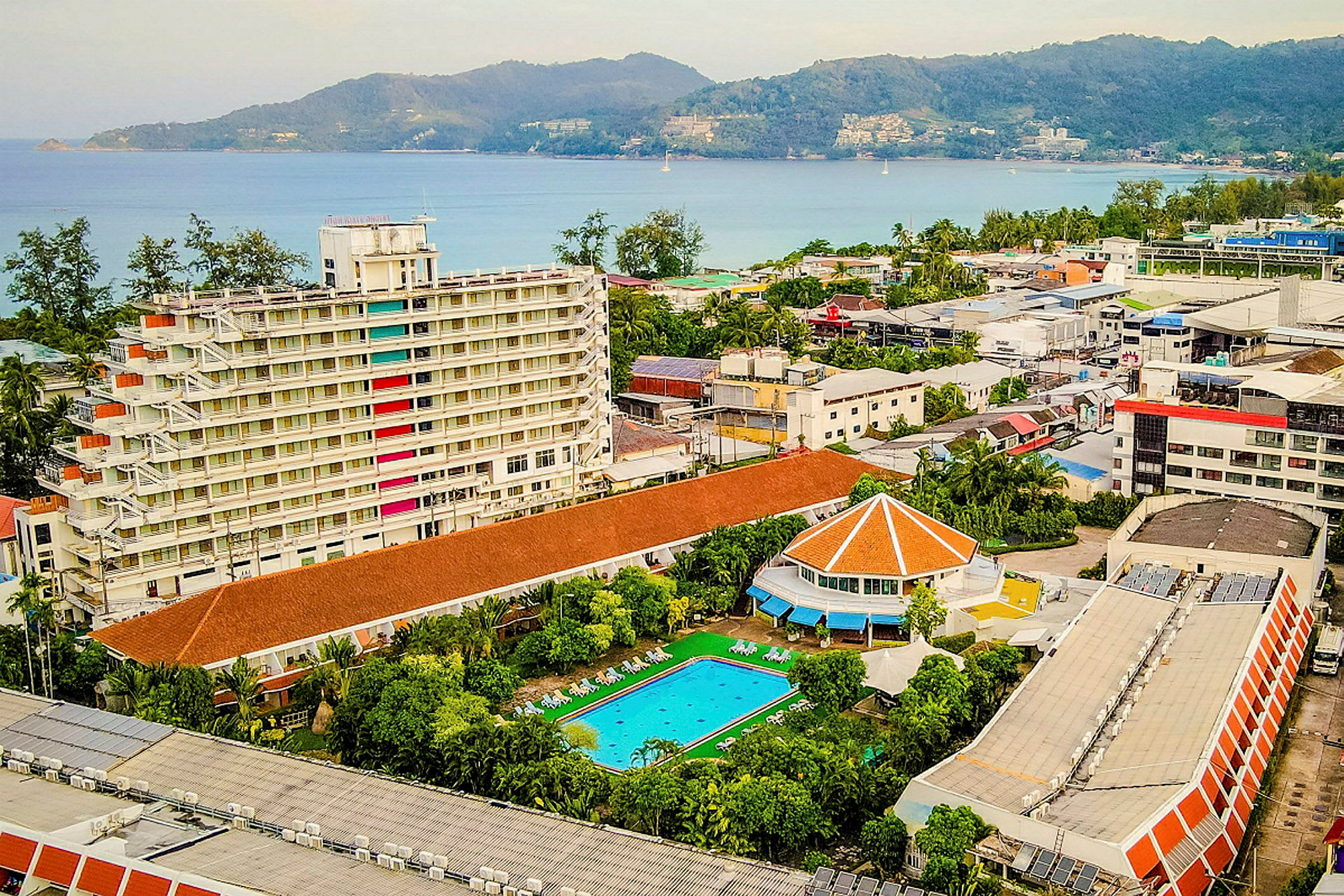 Vy över ett hotellområde med en vit flervåningsbyggnad, en swimmingpool, och en utsikt mot havet. (AI-genererad text)