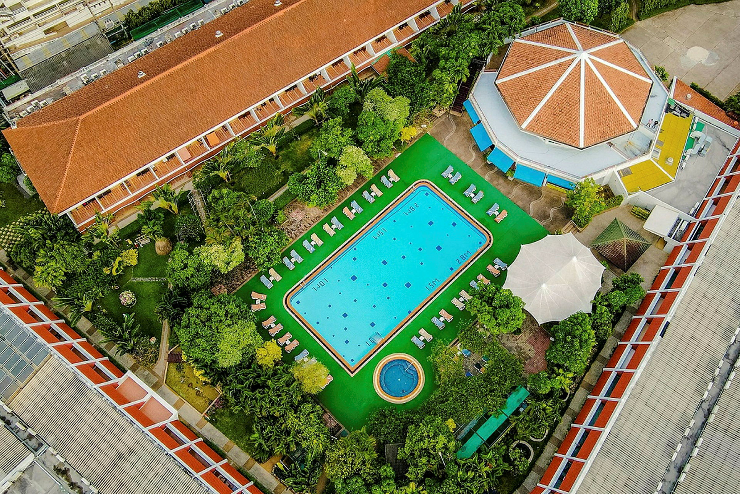 Flygfoto av ett hotellområde med en pool omgiven av solstolar samt grönskande vegetation och byggnader. (AI-genererad text)