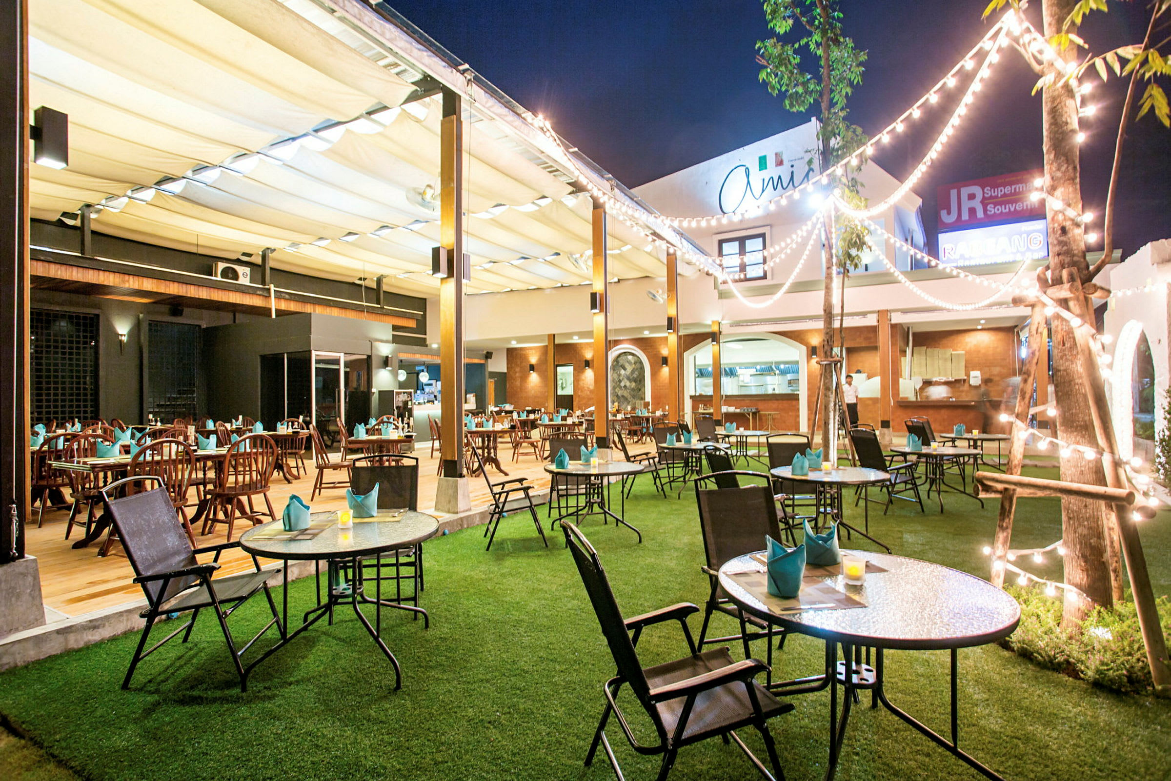 En utomhusrestaurang med dekorerade bord och stolar, gräsmatta och ljusslingor. (AI-genererad text)