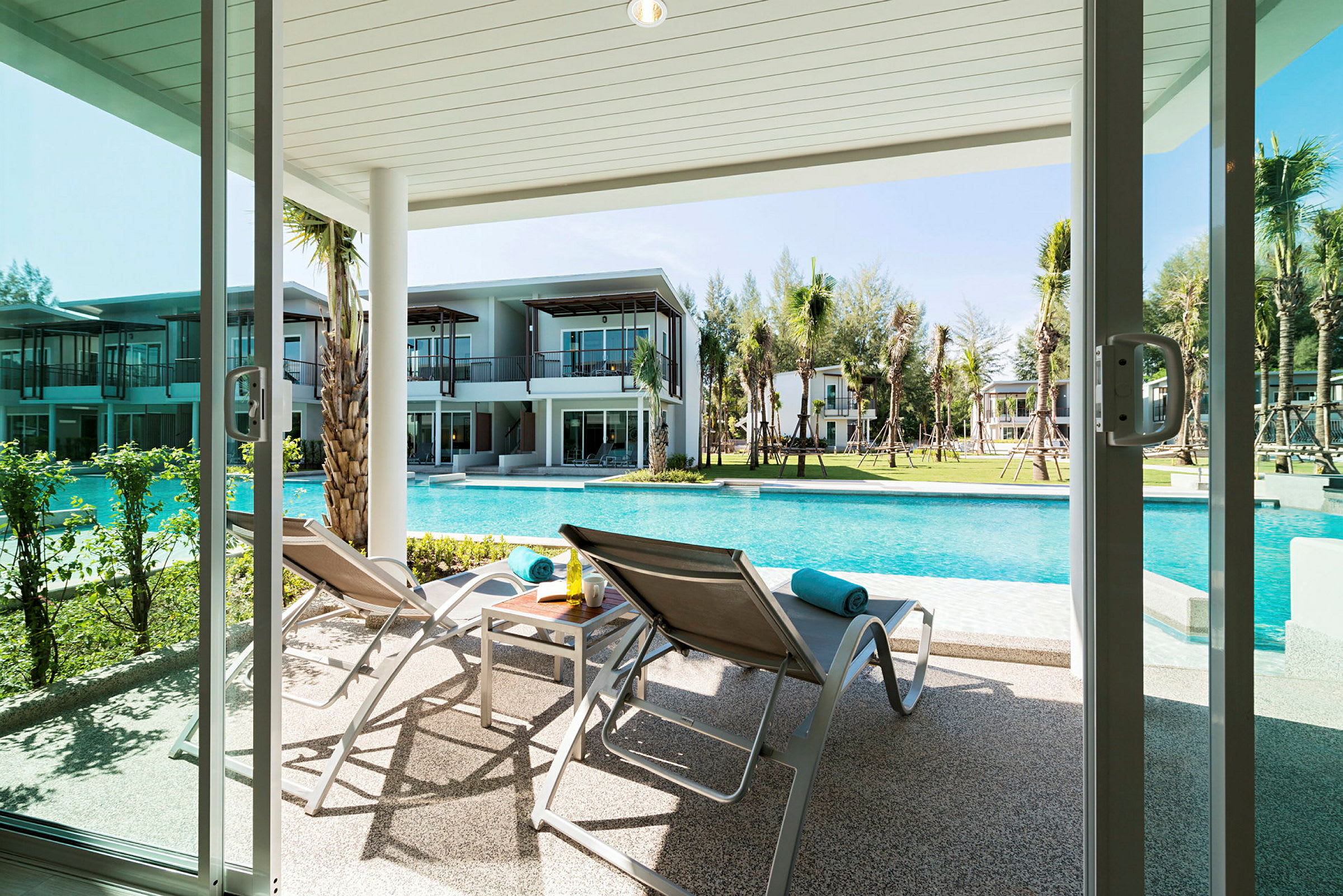 Bild på en uteplats med solstolar och ett bord, vid en pool med moderna byggnader i bakgrunden. (AI-genererad text)