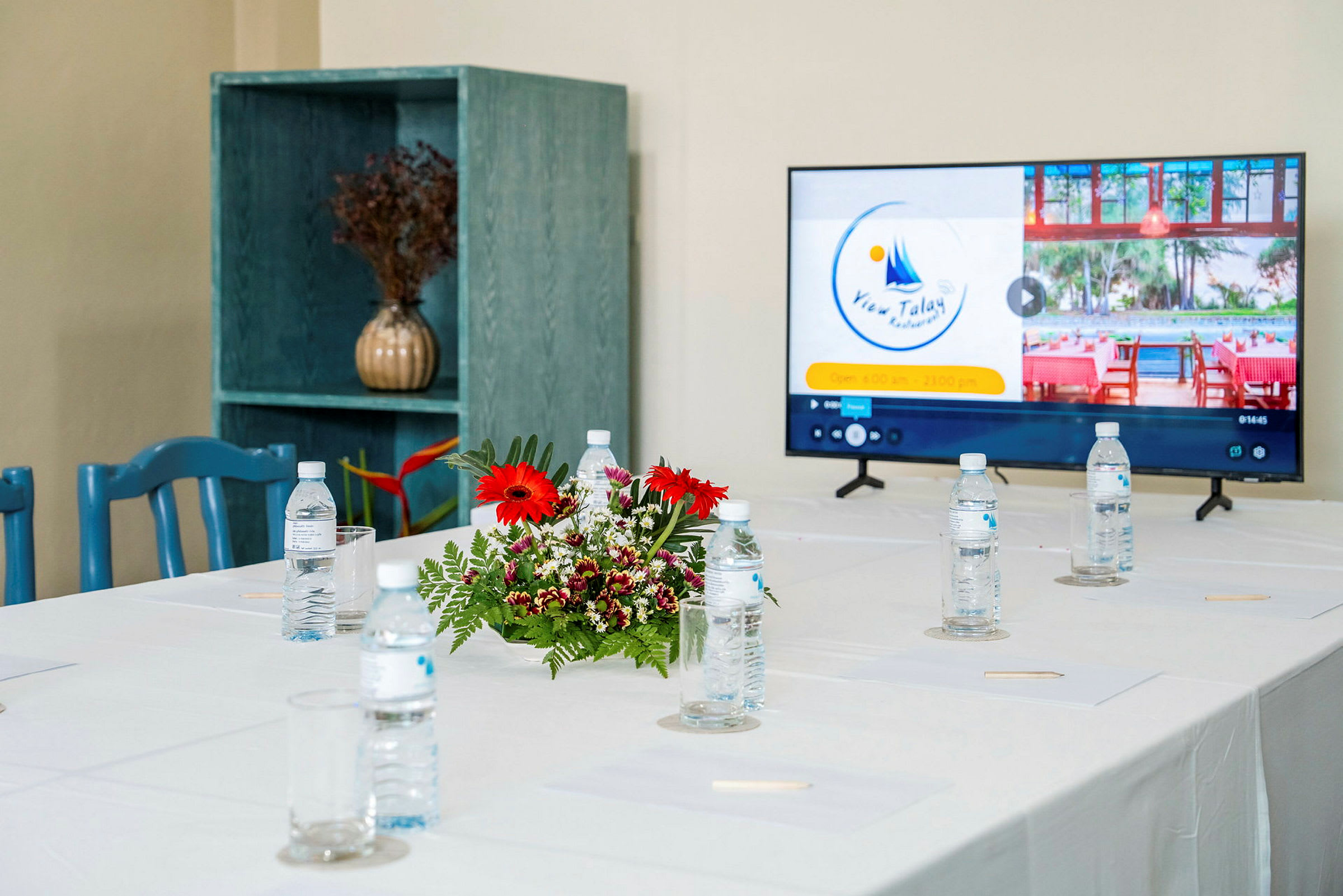 Ett konferensbord med vattenflaskor och en blomsterdekoration, samt en tv-skärm som visar information om View Talay Restaurant. (AI-genererad text)