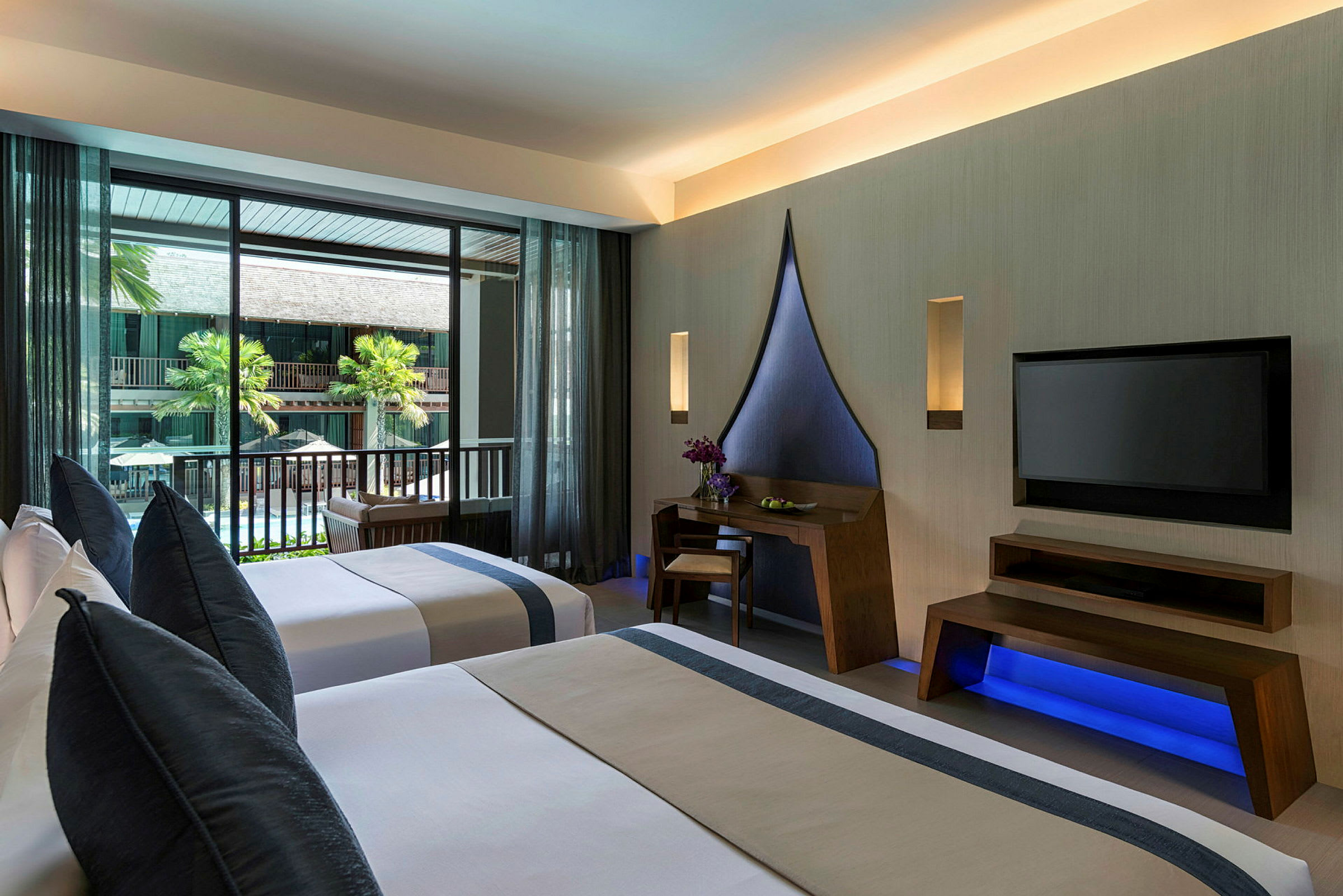 Hotellrum med två enkelsängar, skrivbord och TV. Balkong med utsikt mot en byggnad och palmer. (AI-genererad text)