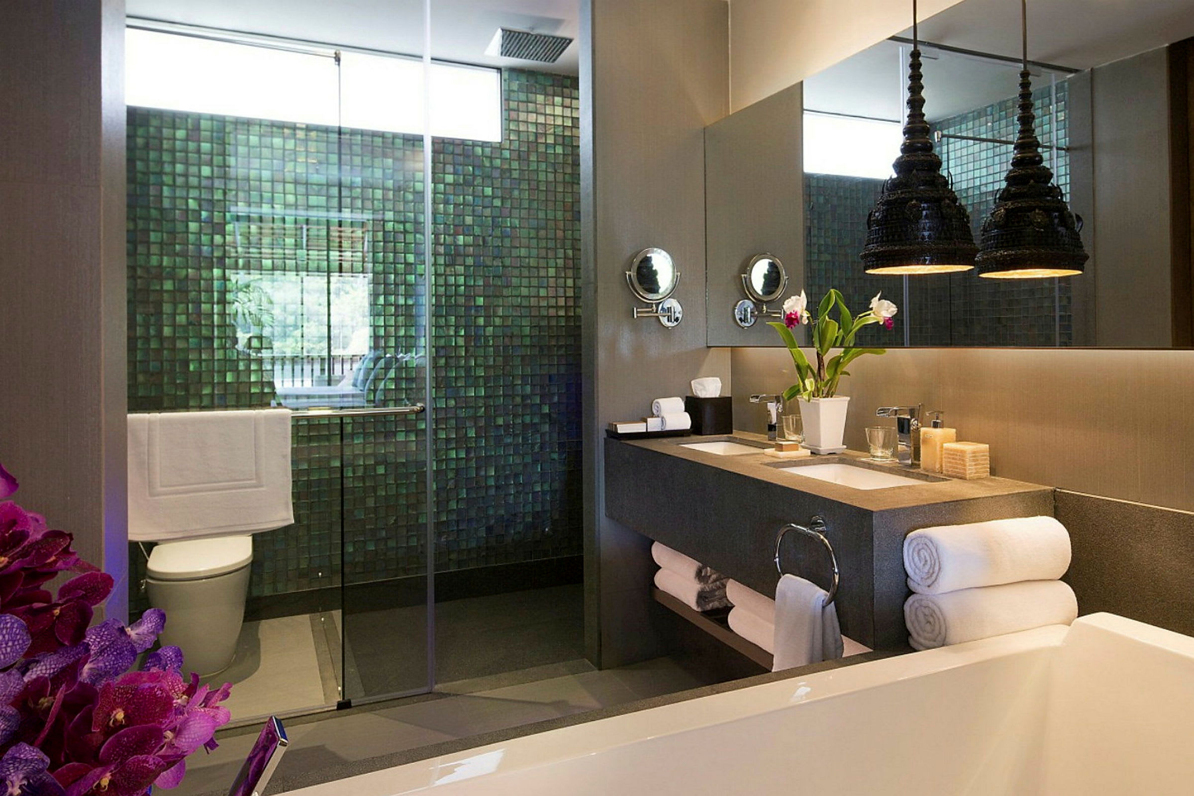 Ett modernt badrum med en dusch med glasdörr, en dubbelhandfat med speglar ovanför, en dekorativ vas med blommor och handdukar prydligt staplade. (AI-genererad text)