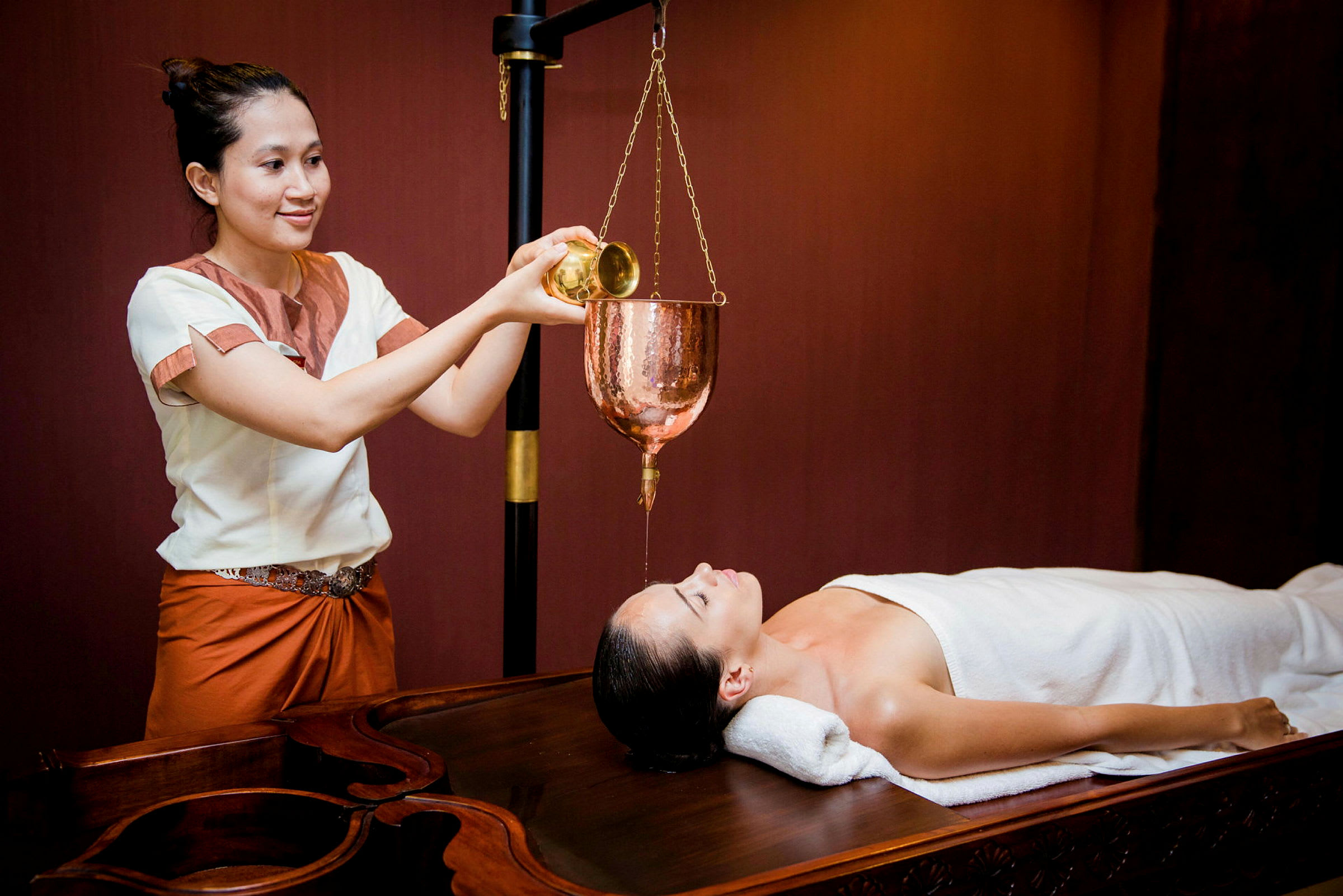 En person utför en spa-behandling med olja hälld från en kopparbehållare på en person som ligger på en massagebänk. (AI-genererad text)
