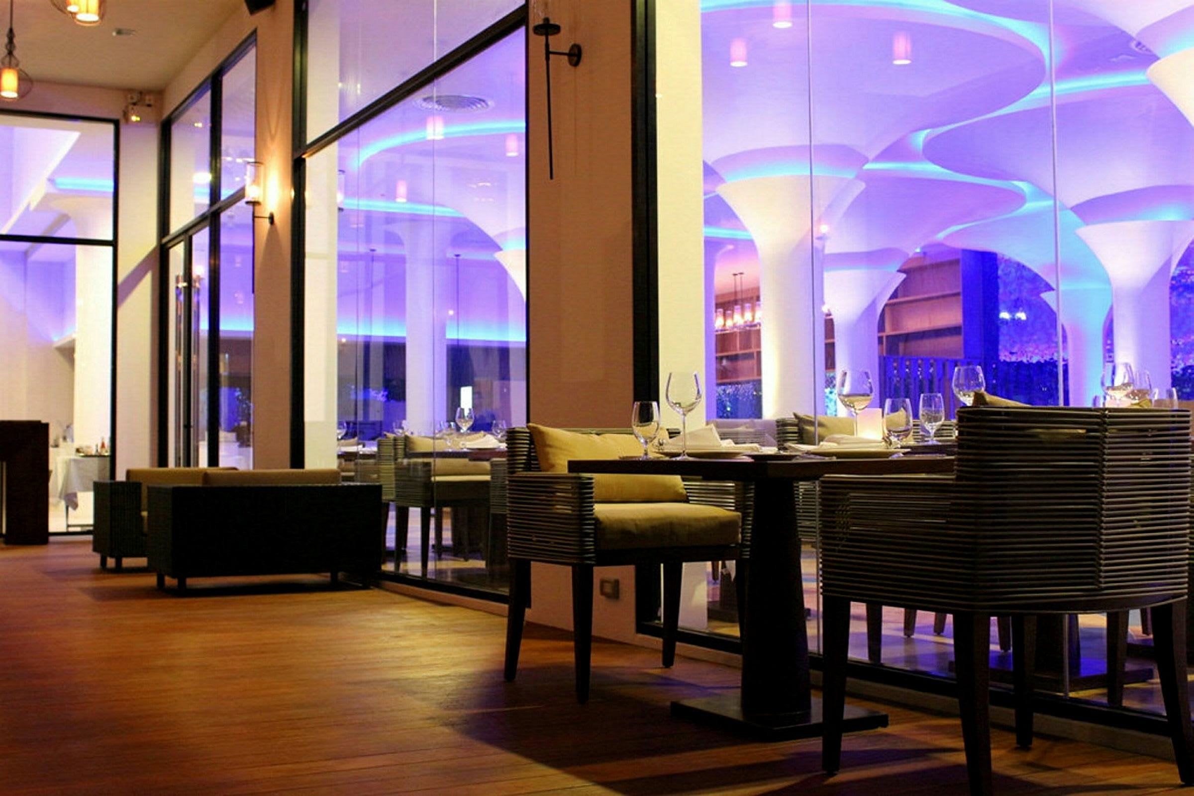 Interiör av en elegant restaurang med glasväggar och moderna möbler, belysning i blått och varmt ljus. (AI-genererad text)