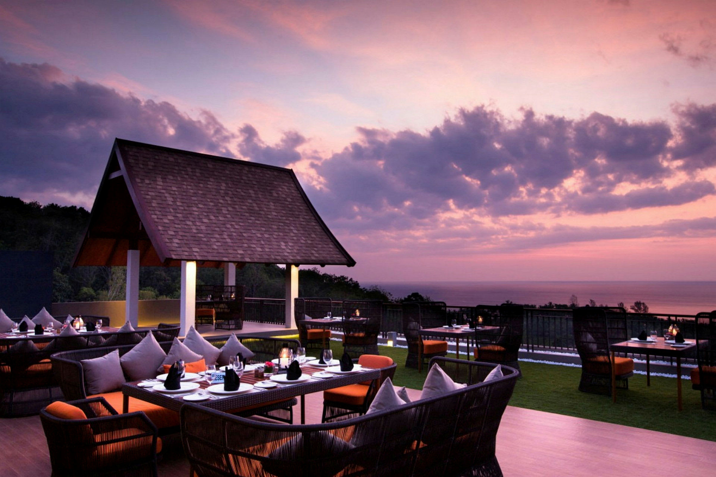 En utomhusrestaurang med bord och stolar under en lila och rosa himmel i solnedgången, med utsikt över havet. (AI-genererad text)