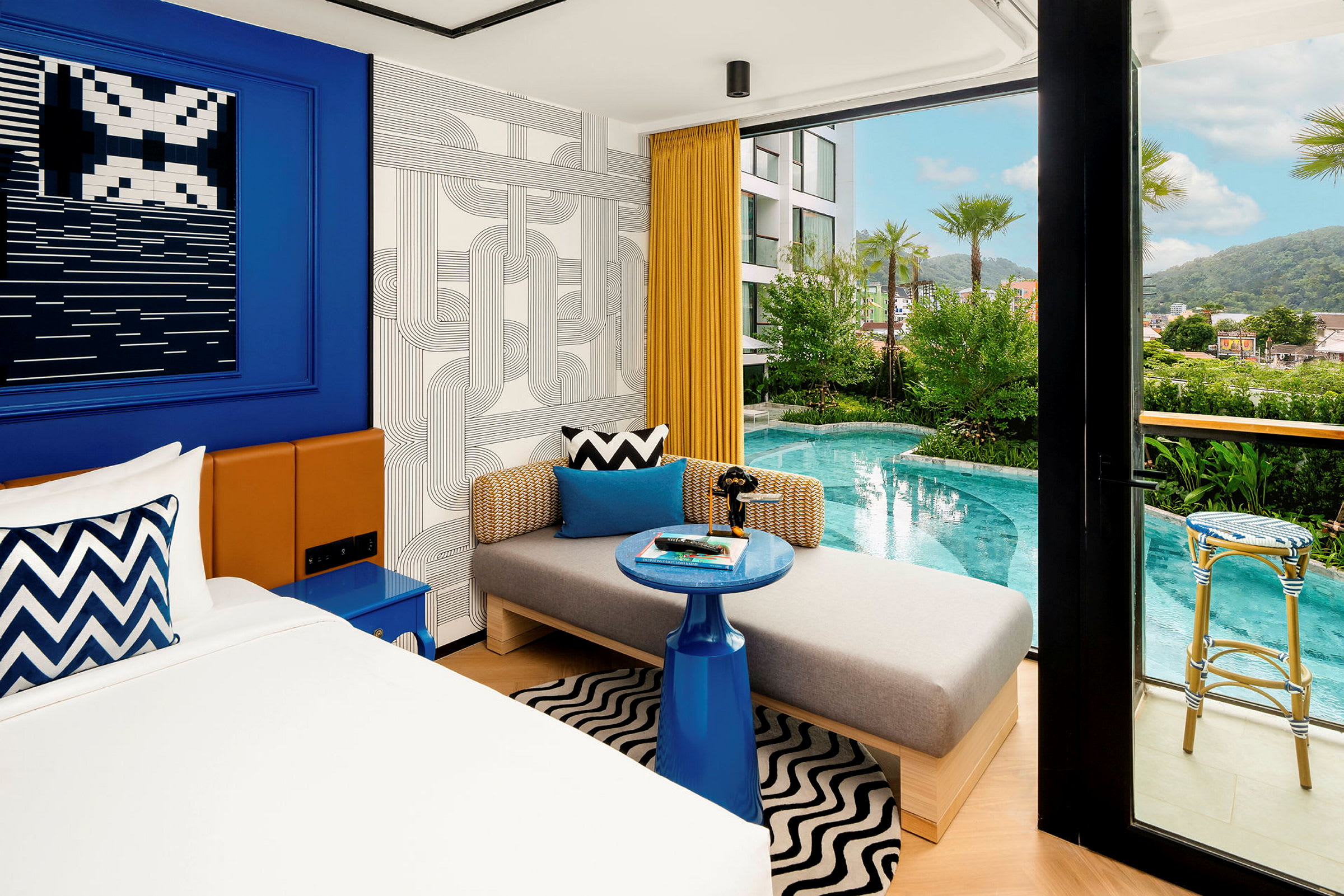 Ett modernt hotellrum med en säng, en soffa och en liten blå sidobord. Utanför finns en pool och omgivande grönska. (AI-genererad text)