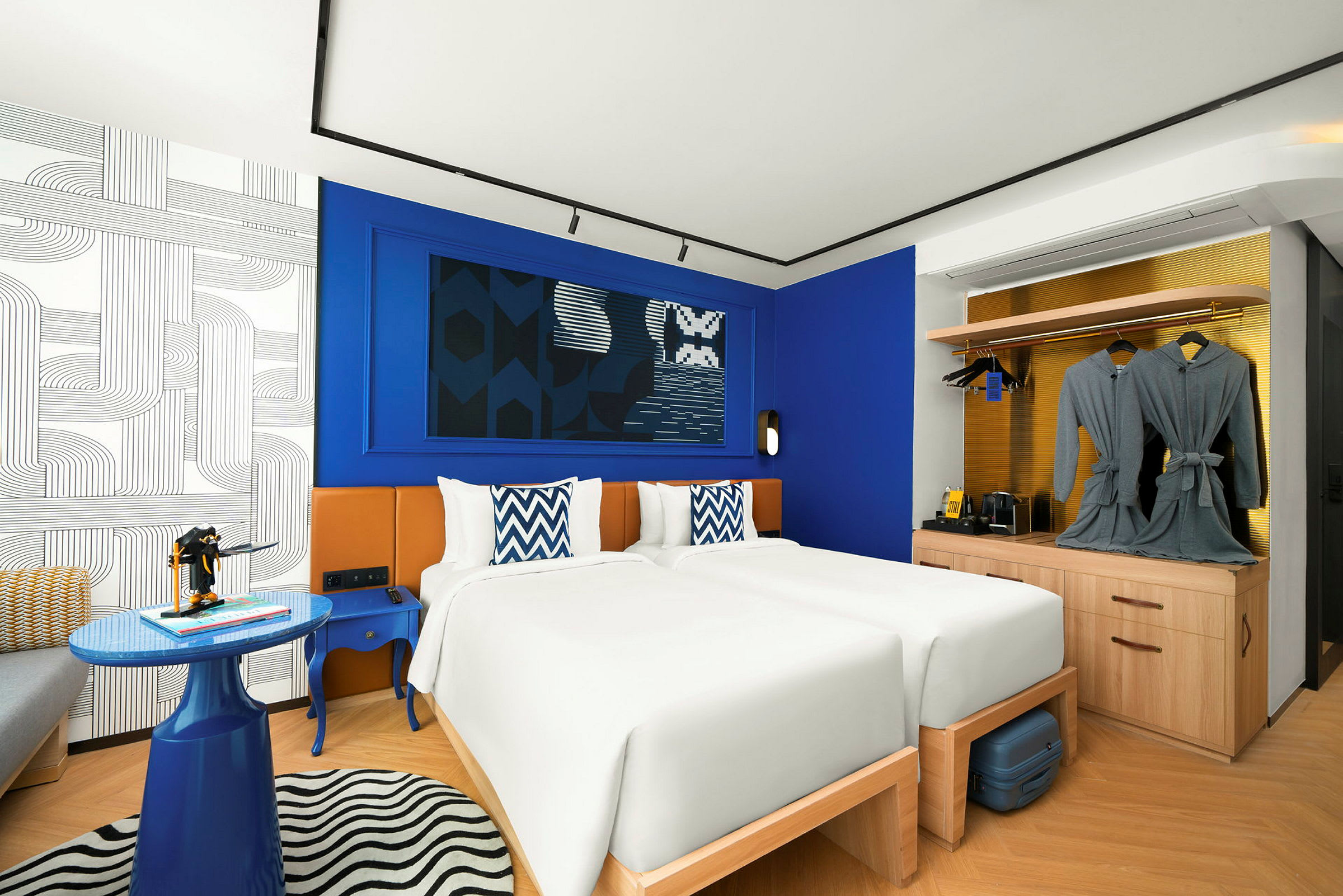 Ett hotellrum med två enkelsängar, blå och vita dekorativa kuddar, moderna möbler och en garderob med morgonrockar. (AI-genererad text)
