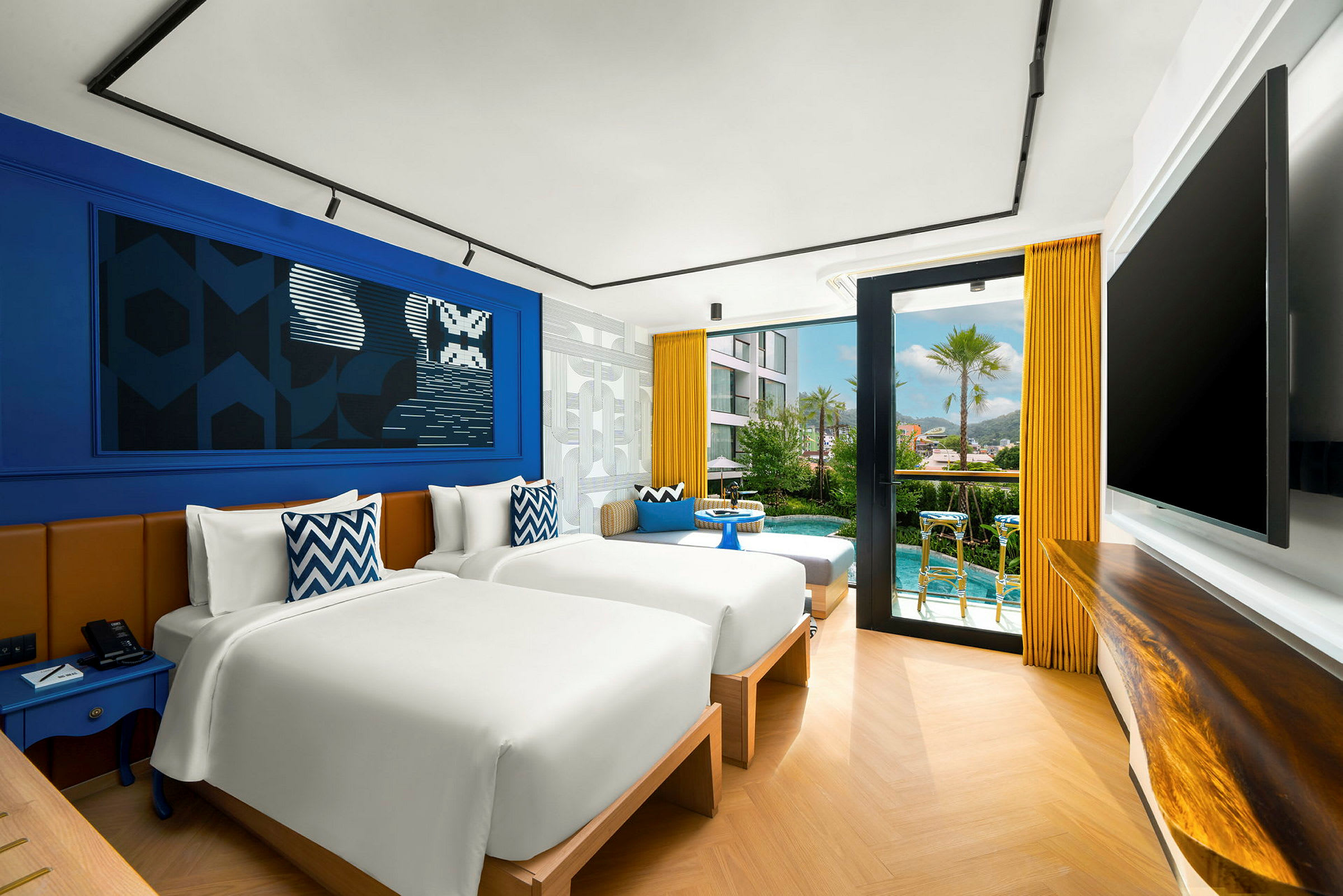 Ett modernt hotellrum med två separata sängar, blå och gula inredningsdetaljer samt en glasdörr som leder ut till en liten terrass och poolområde. (AI-genererad text)