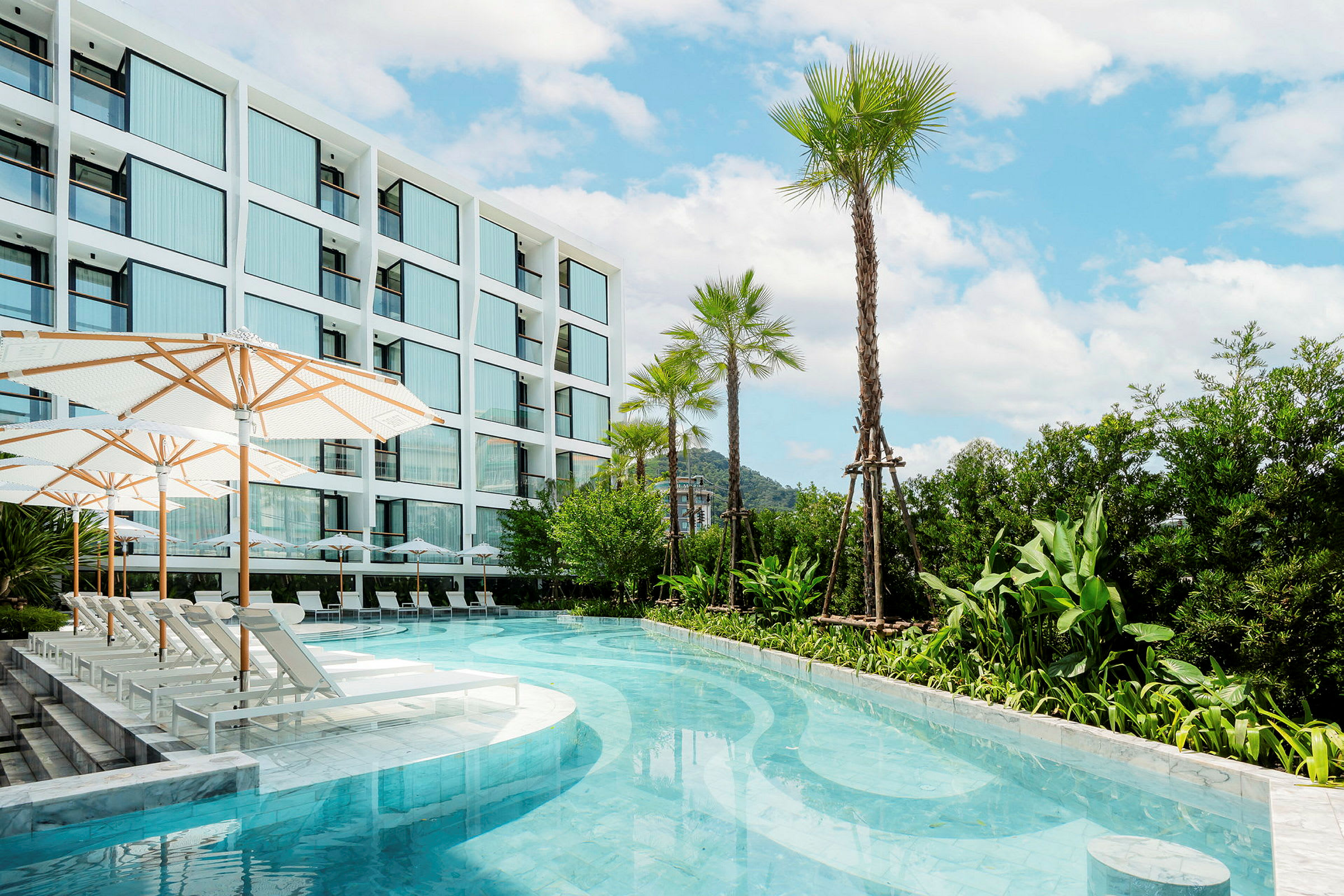 En utomhuspool med solstolar och parasoller ligger intill en modern hotellbyggnad omgivet av gröna växter och palmer under en blå himmel. (AI-genererad text)