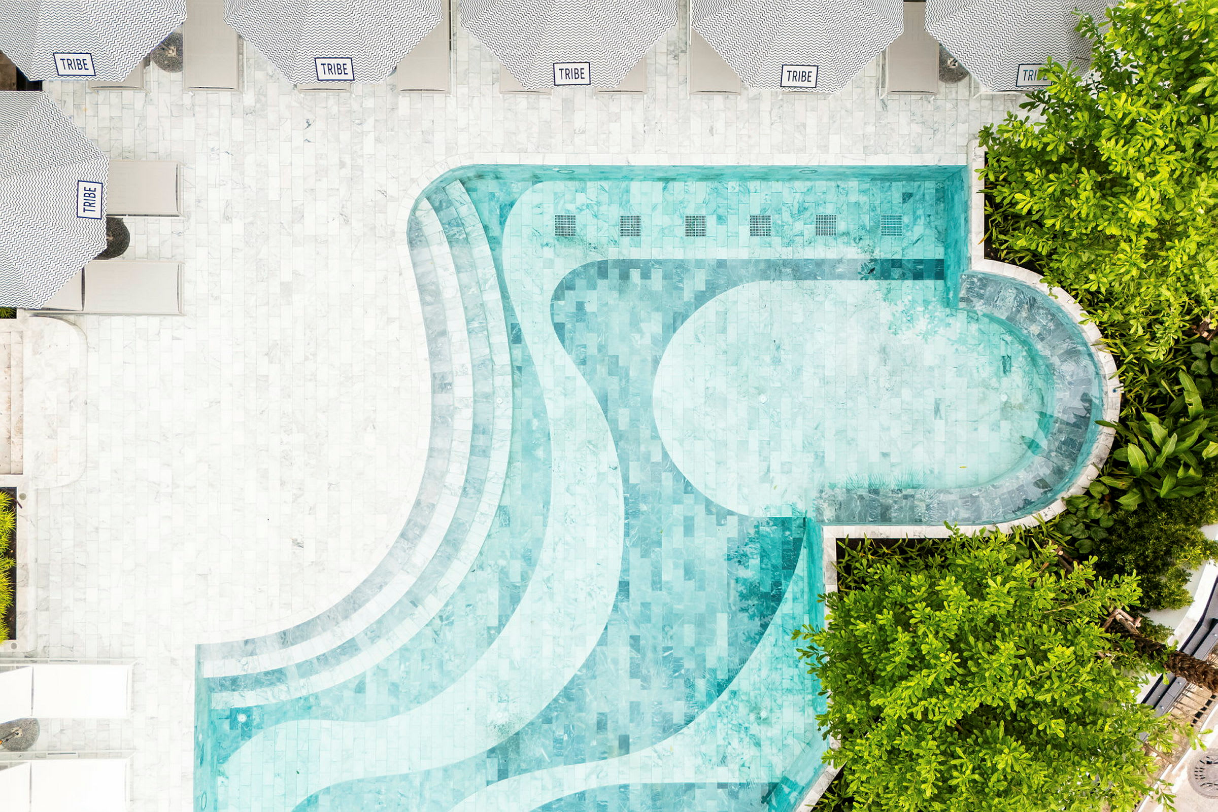 En flygbild av en pool med ljusblått vatten omgiven av trädkant och solstolar under parasoll. (AI-genererad text)