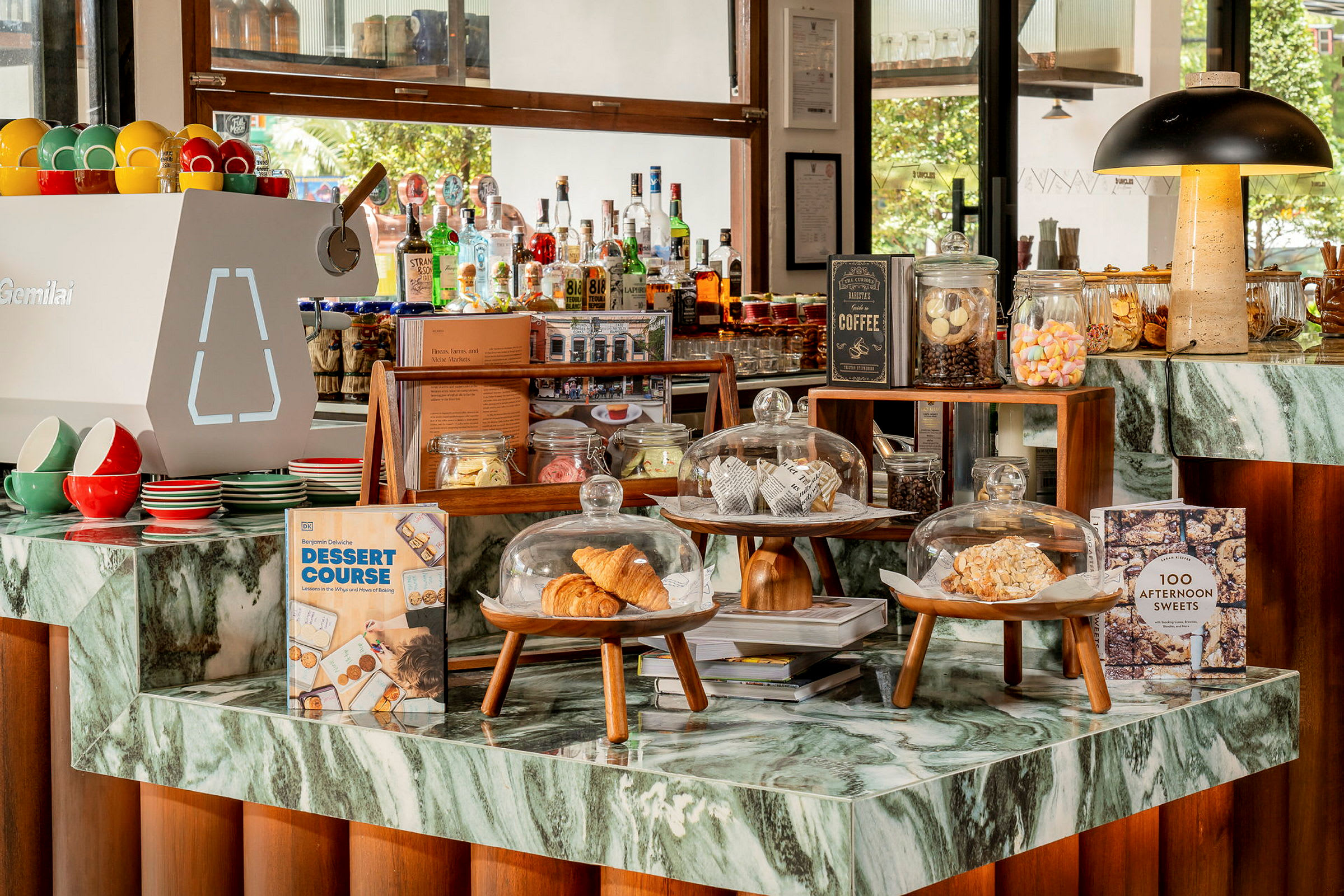 Ett kafé med en marmordisk där croissanter och andra bakverk presenteras under glaskupor. Det finns även färgglada koppar, böcker och burkar med godis på disken. (AI-genererad text)