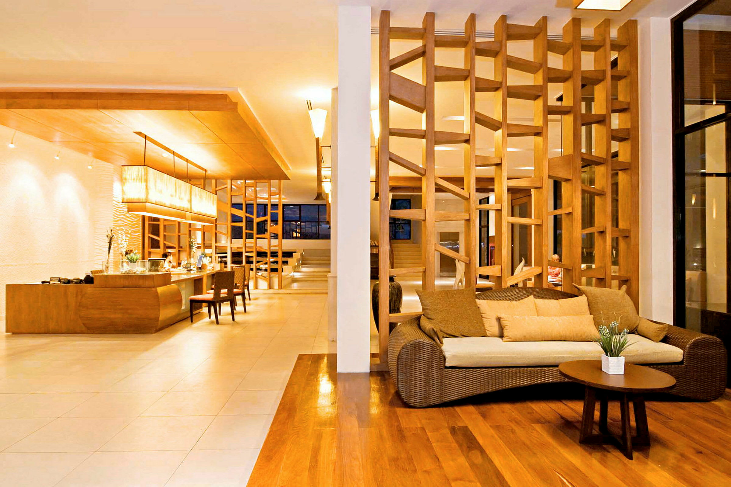 En hotellobby med trägolv, en soffa med kuddar och ett lågt bord i förgrunden, samt ett designat träparti som avdelning. Bakgrund visar receptionsområdet med belysning ovanför. (AI-genererad text)