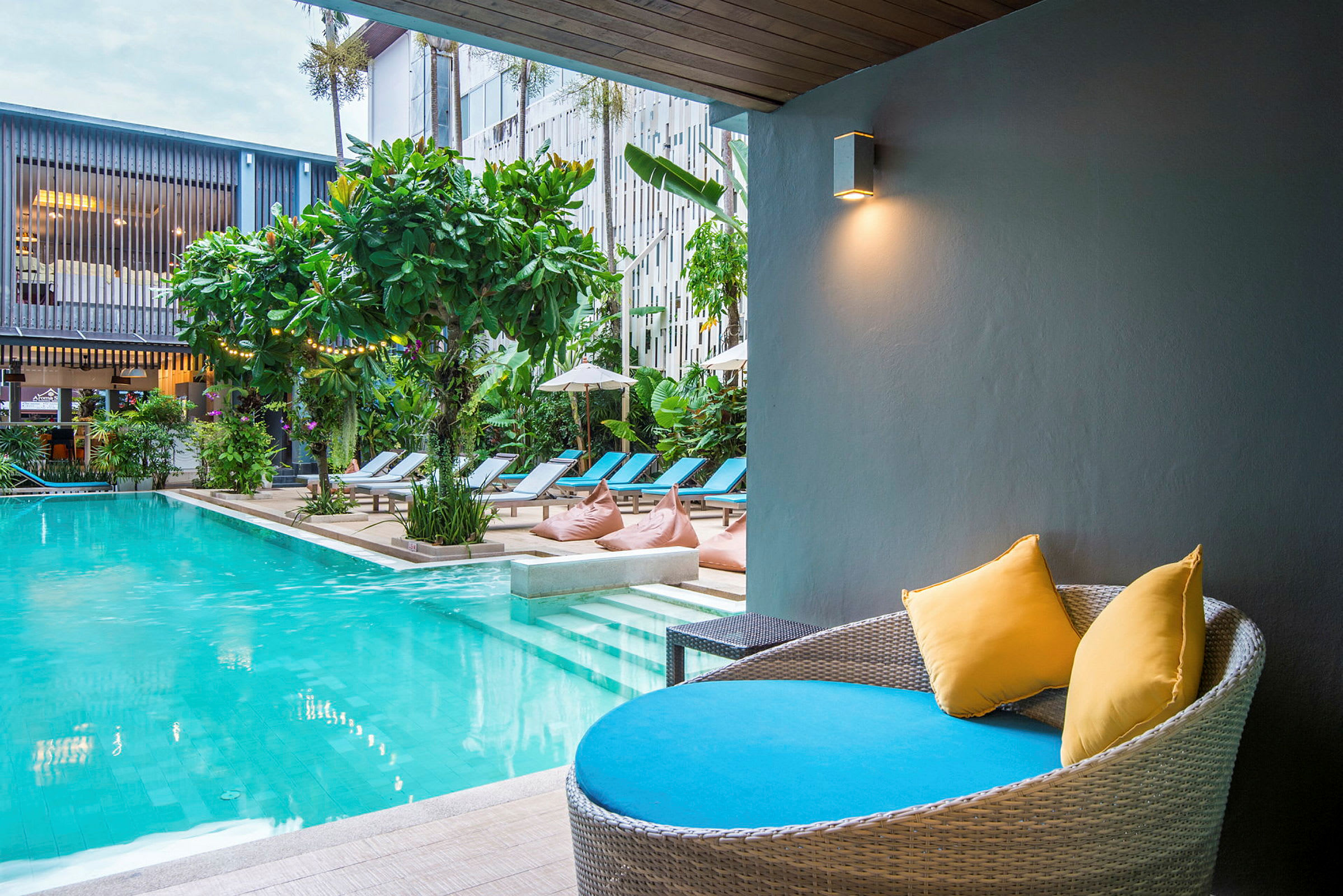 Modern pool med solstolar och växtlighet. En sittplats med kuddar i förgrunden. (AI-genererad text)