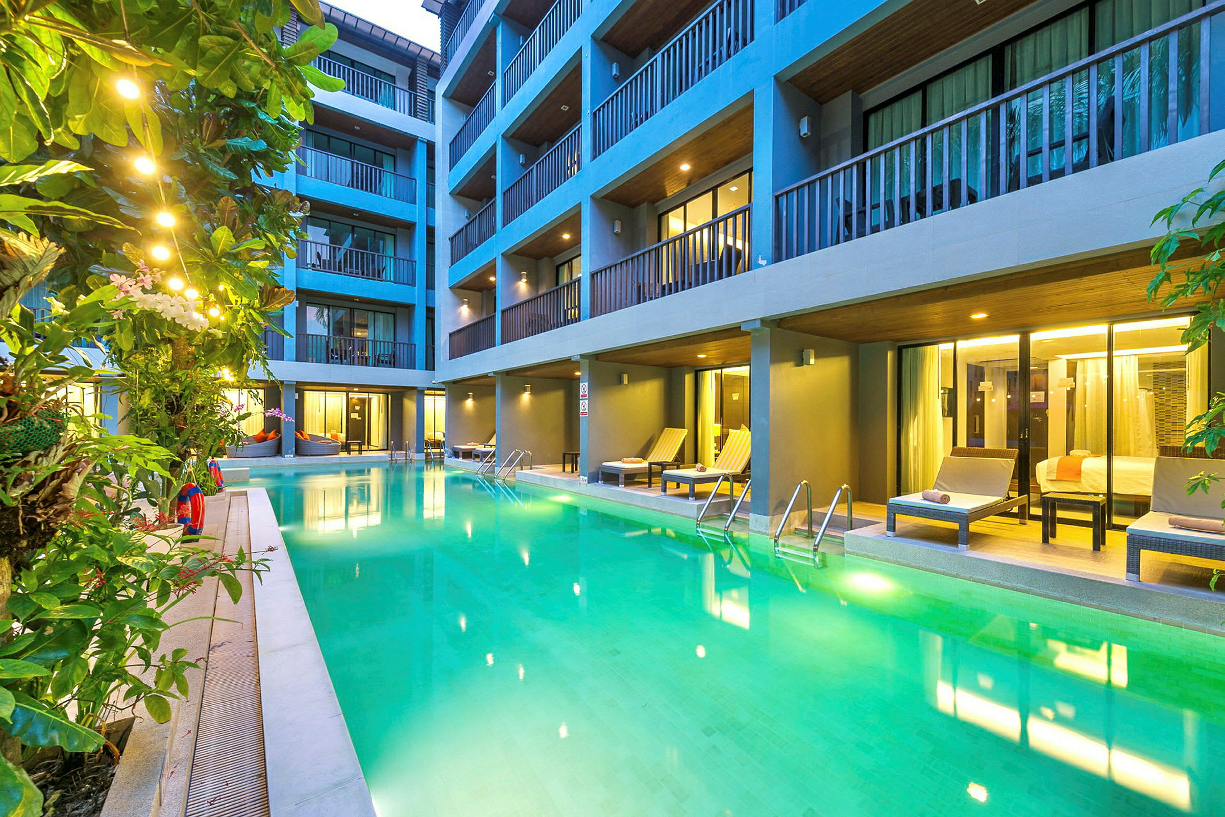 Pool med solstolar och kringliggande växter framför hotellrum. (AI-genererad text)