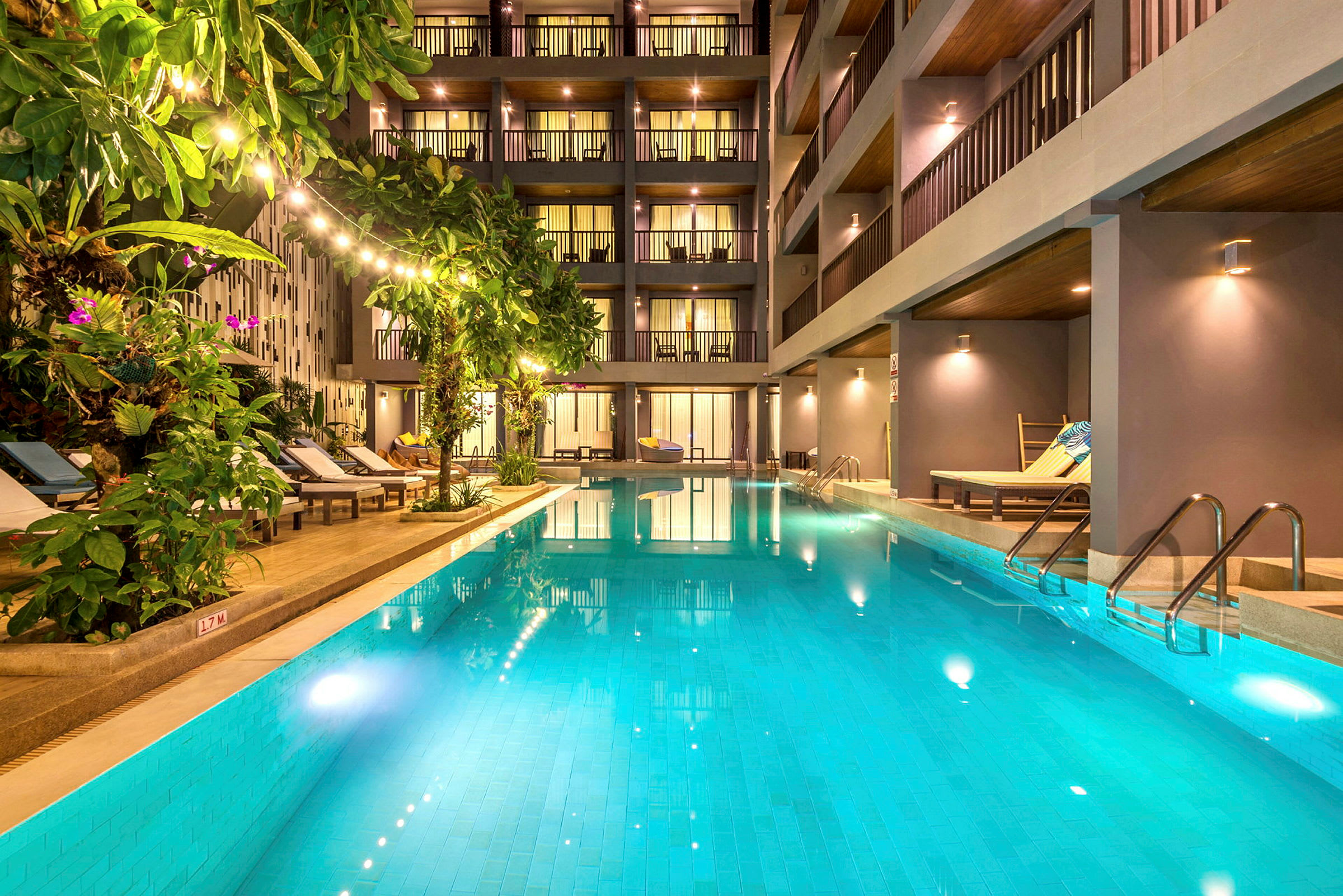 En utomhuspool omgiven av solstolar och grönska framför ett modernt hotell. (AI-genererad text)
