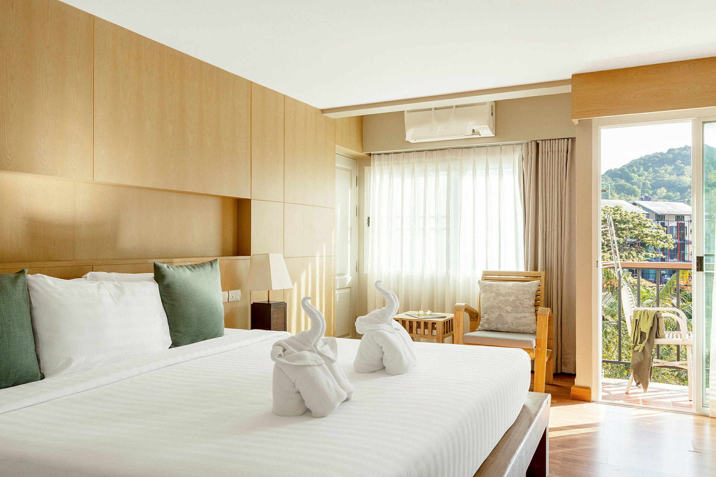 Ett välupplyst hotellrum med en stor säng täckt av vita lakan och kuddar. På sängen finns två handdukar vikta till formen av svanar. Rummet har en träinredning, en sittgrupp med kuddar och en balkong med utsikt mot grönska och byggnader. (AI-genererad text)