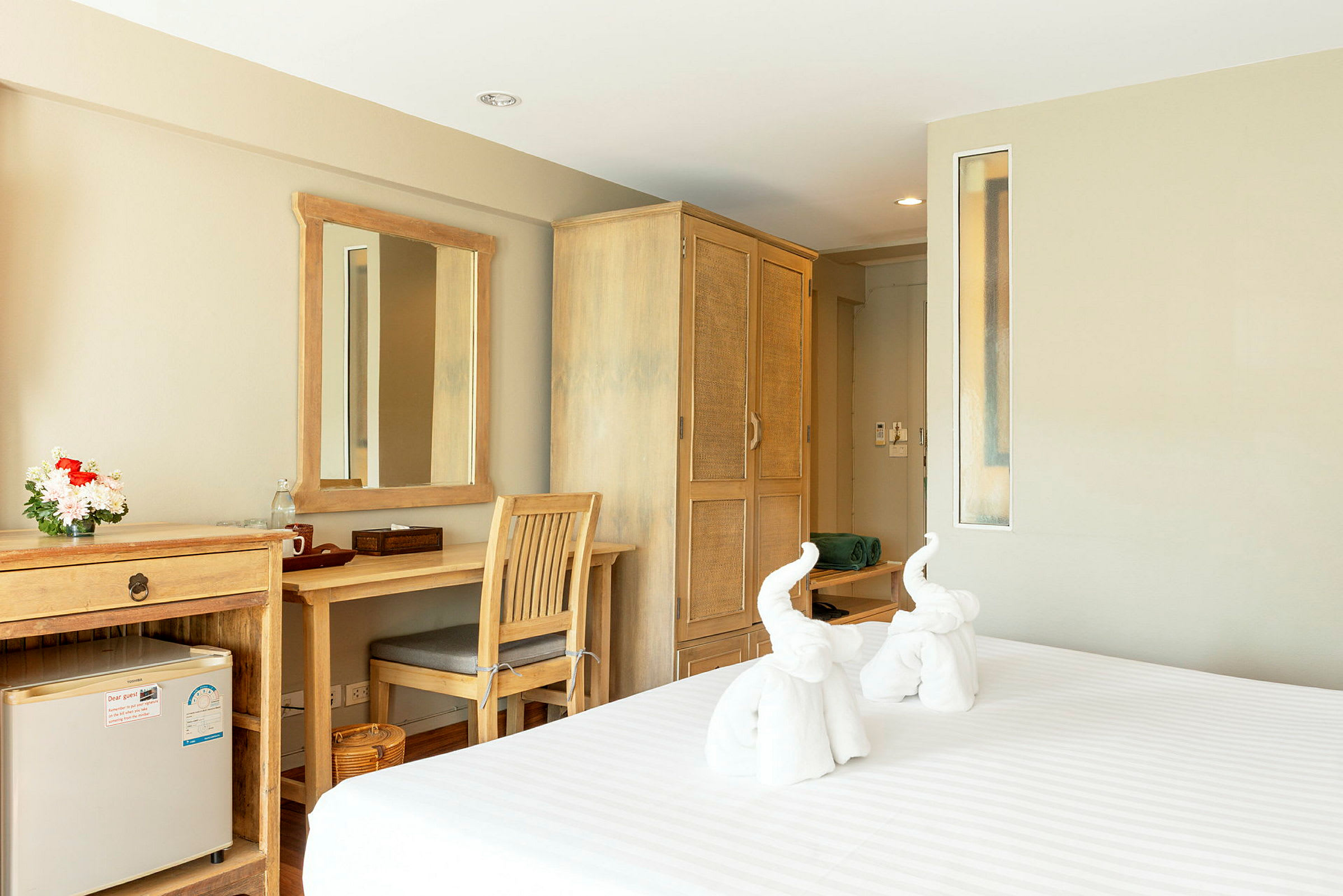 Ett hotellrum med träinredning, en spegel ovanför ett skrivbord och ett kylskåp. En säng med vikta handdukar i form av elefanter. (AI-genererad text)