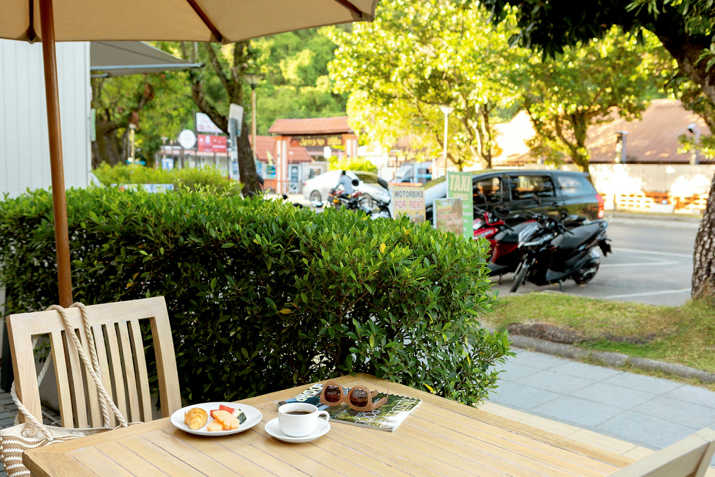 Ett bord utomhus med ett parasoll och en tallrik med frukost, kaffe och tidningar. Bakom bordet finns buskar och en gata med motorcyklar och bilar. (AI-genererad text)