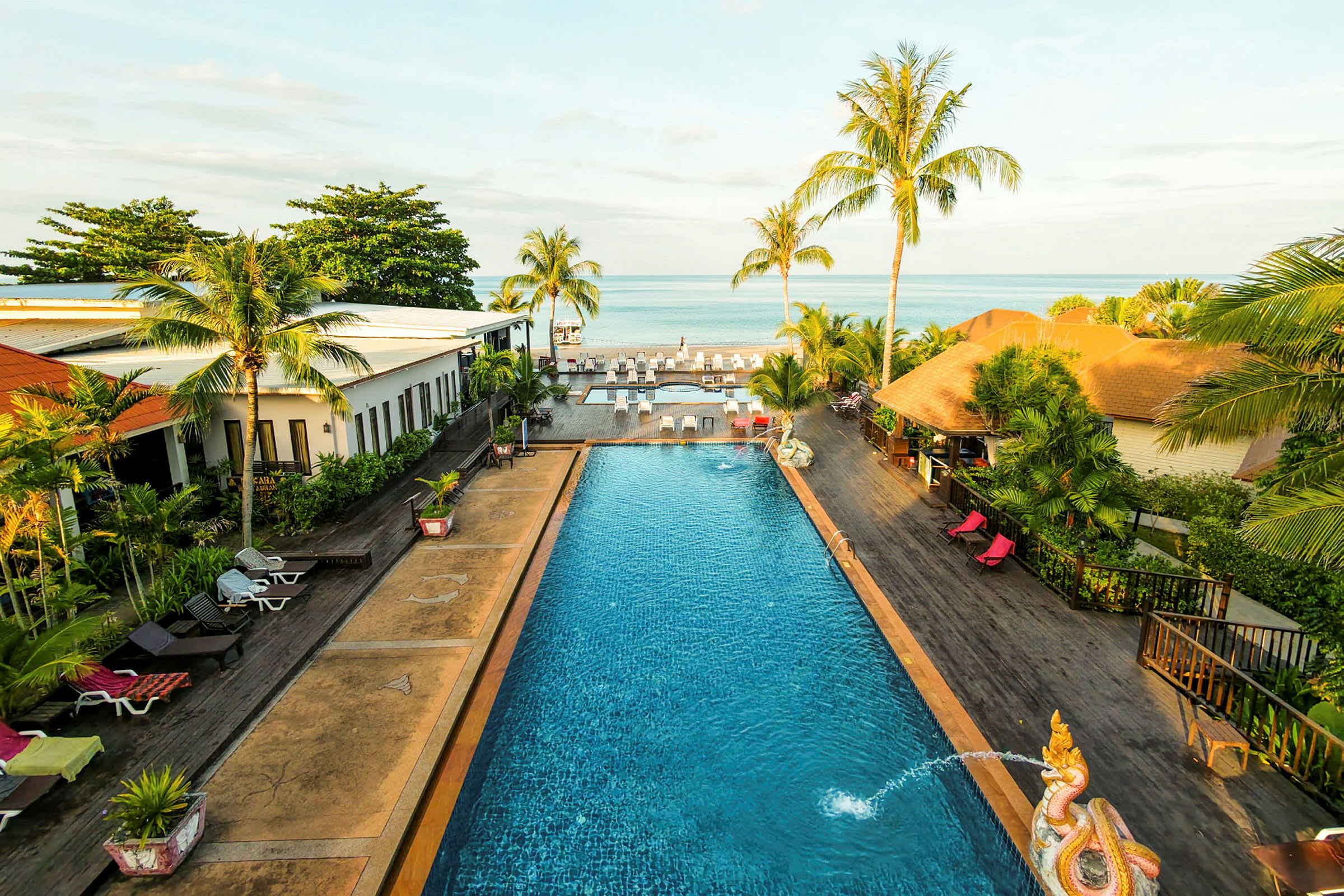 Ett hotellområde med en pool omgiven av byggnader, palmer och solstolar, med havet i bakgrunden. (AI-genererad text)