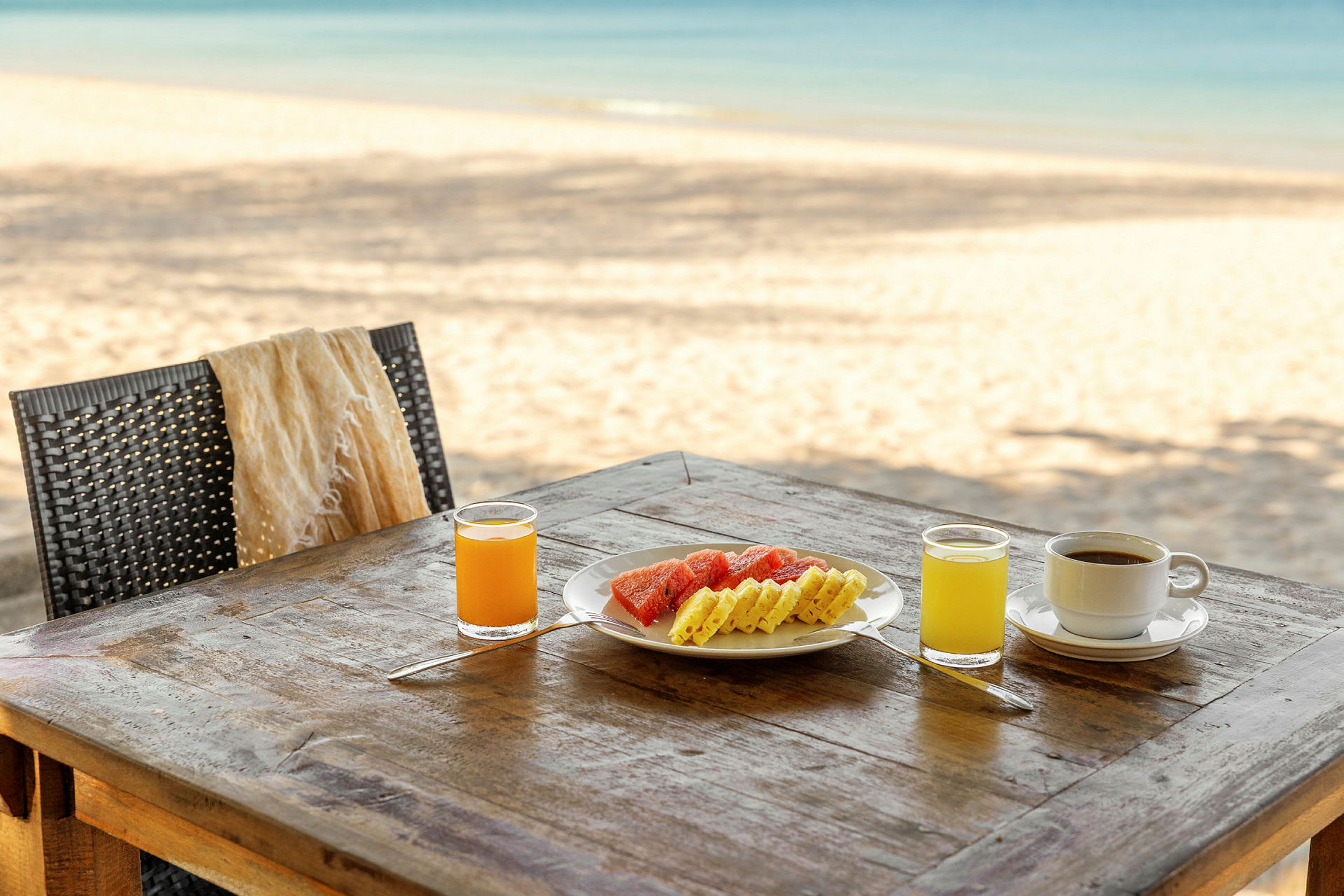 Ett bord på stranden med ett fruktfat, en kopp kaffe och två glas juice. (AI-genererad text)