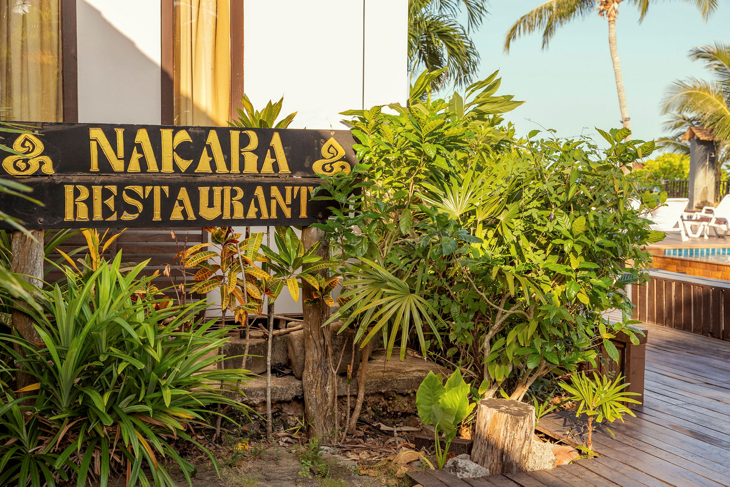 En skylt med texten "Nakara Restaurant" omgiven av grönska, med en träplattform och en pool i bakgrunden. (AI-genererad text)