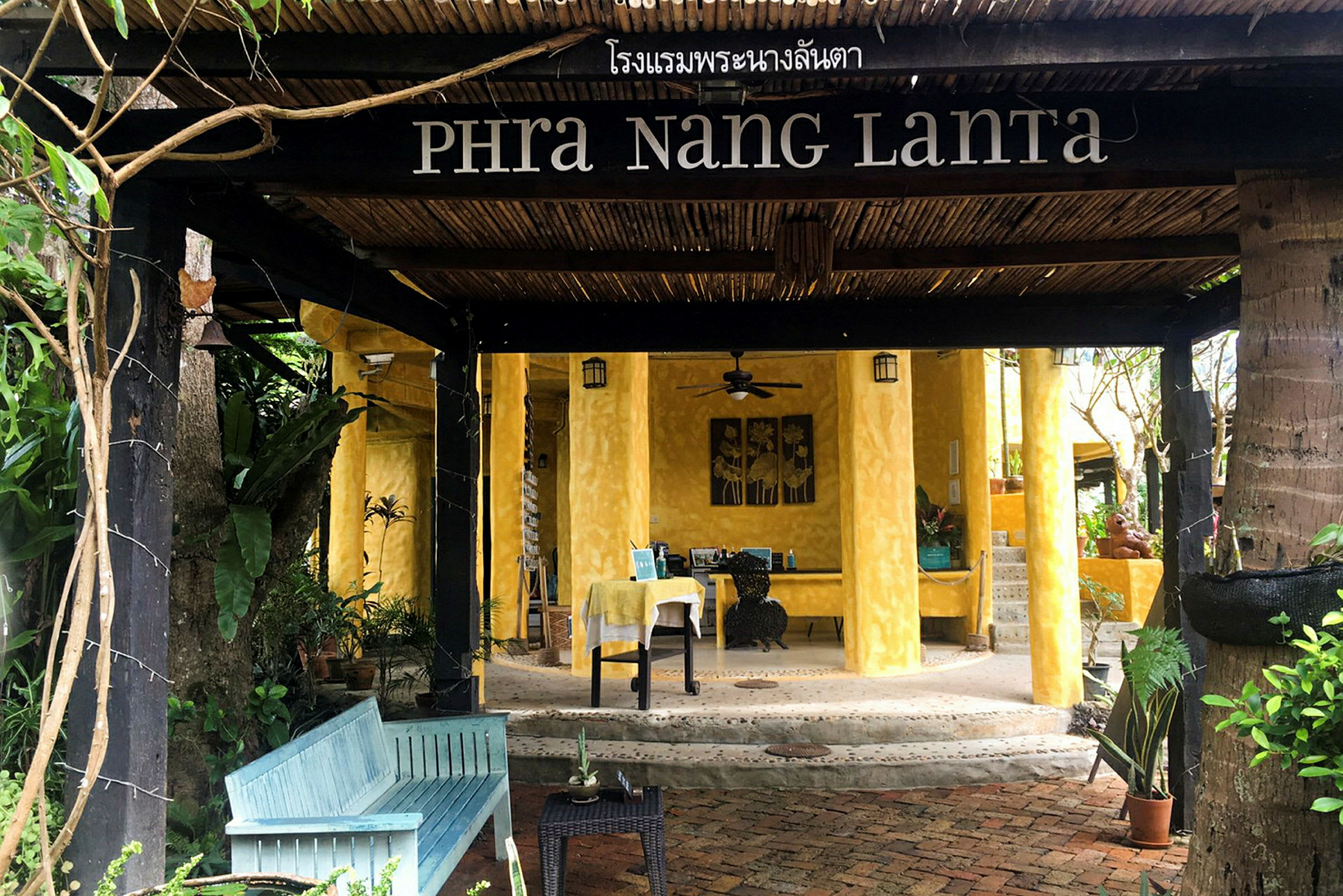 Entrén till ett hotell med namnet Phra Nang Lanta, med gula väggar och trädetaljer, omgiven av grönska. (AI-genererad text)