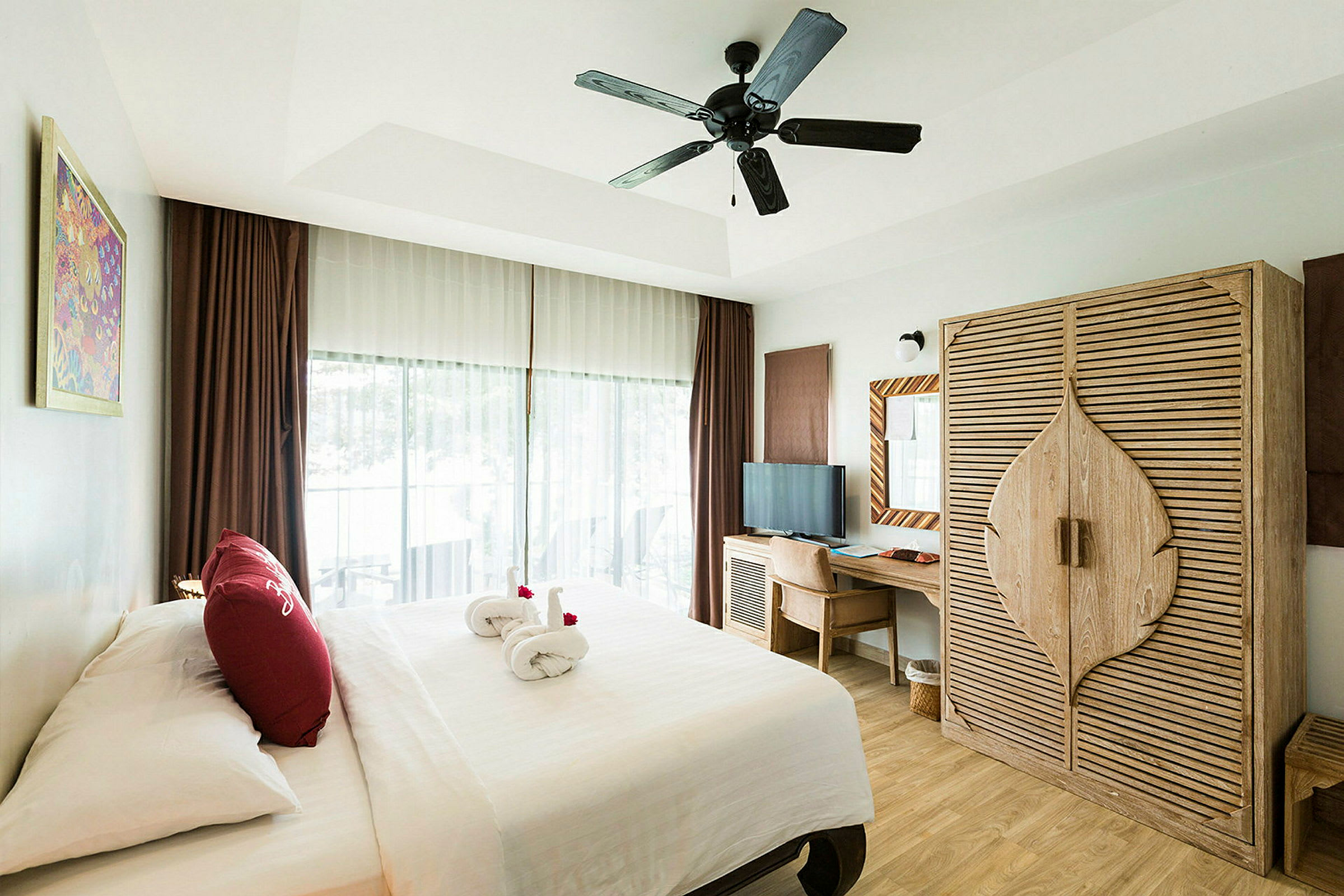 Ett hotellrum med en dubbelsäng, en garderob av trä, en skrivbord med stol och en takfläkt. Ett fönster med gardiner visar en balkong. (AI-genererad text)