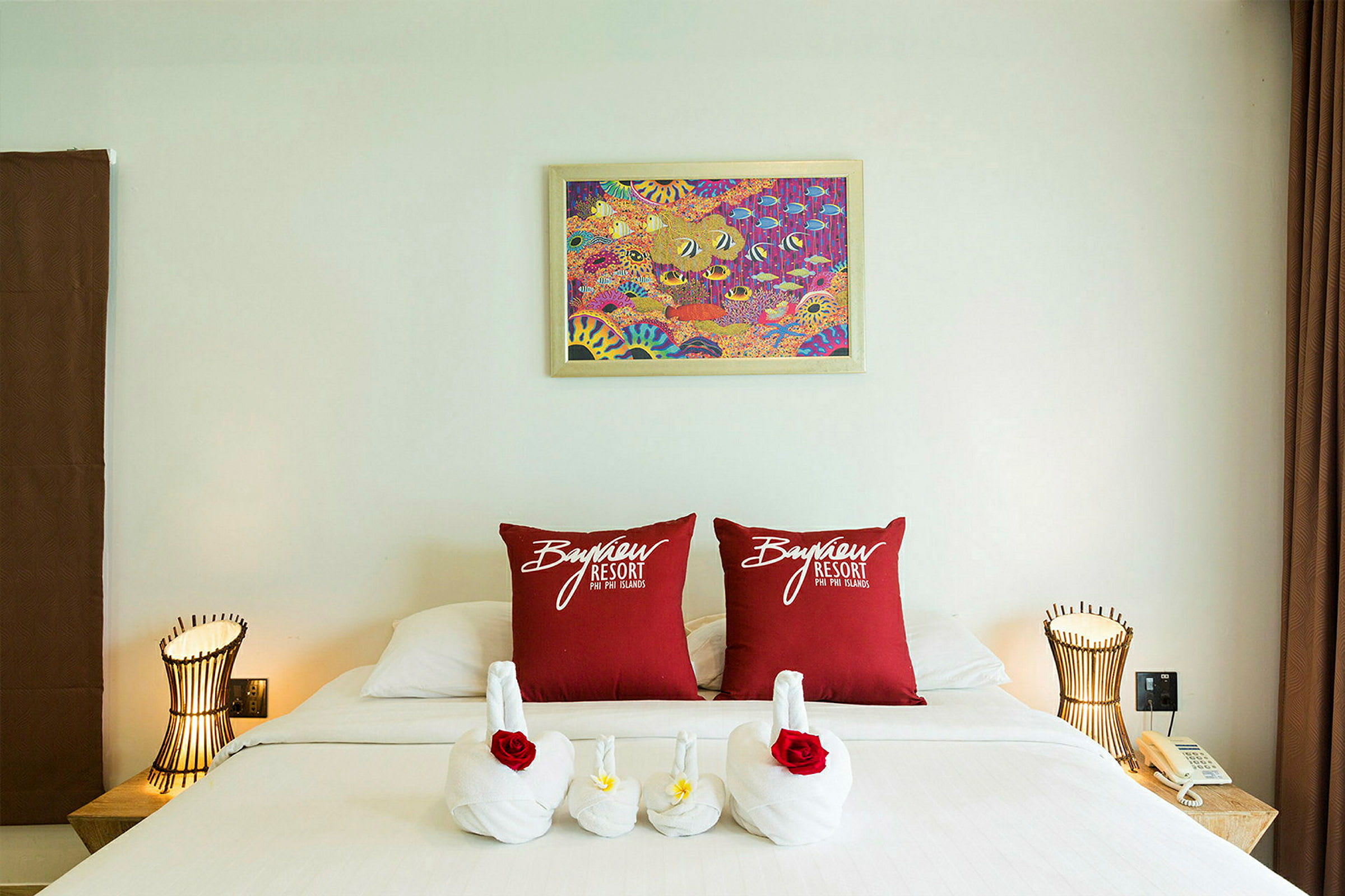 Ett hotellrum med en dekorerad säng. Två röda kuddar med texten ”Bayview Resort” är placerade på sängen, och handdukar är arrangerade som svanar med blommor. Det finns en lampa och en telefon på ett nattduksbord. (AI-genererad text)