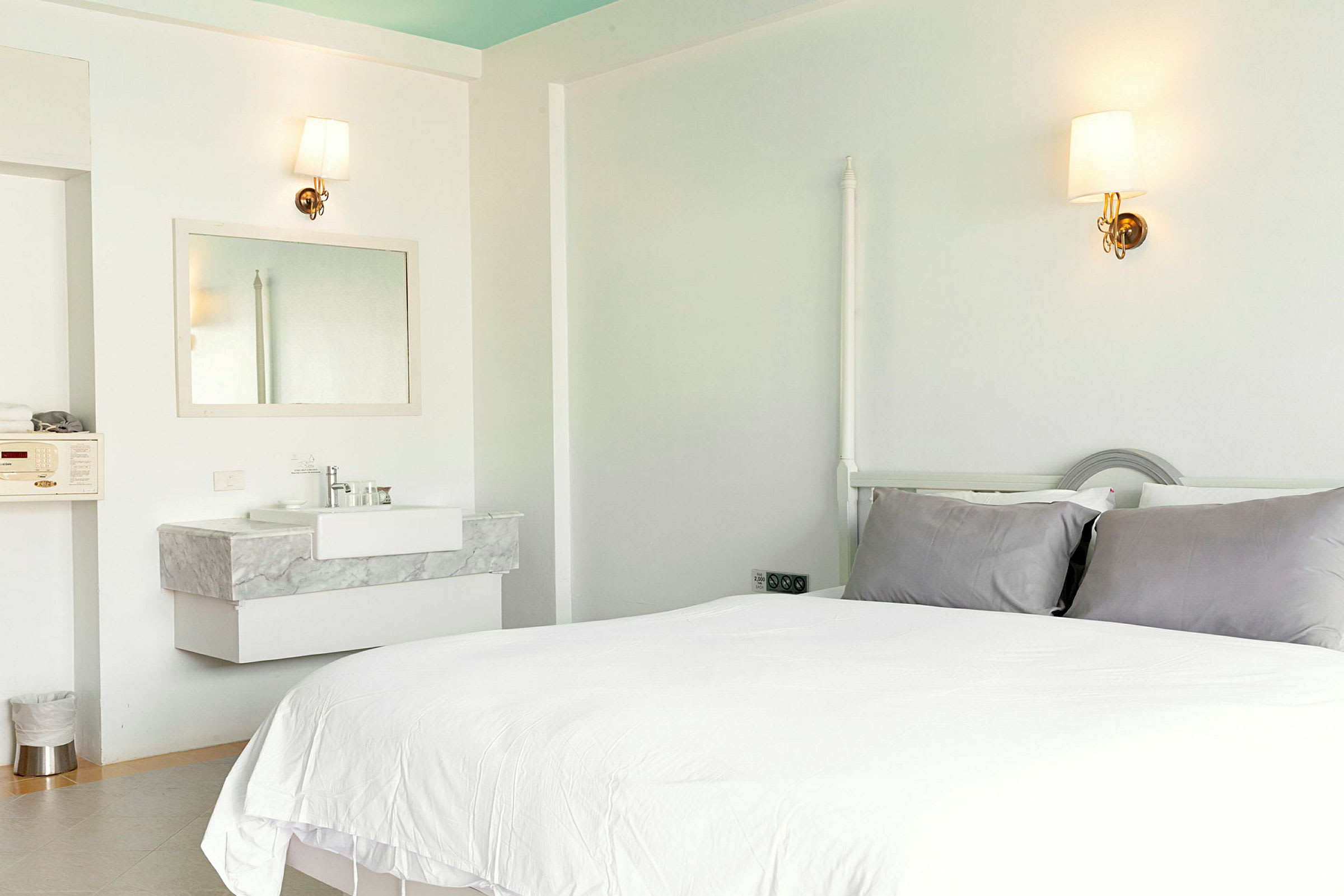 Ett ljust hotellrum med en säng, gråa kuddar, och ett handfat med en spegel. (AI-genererad text)
