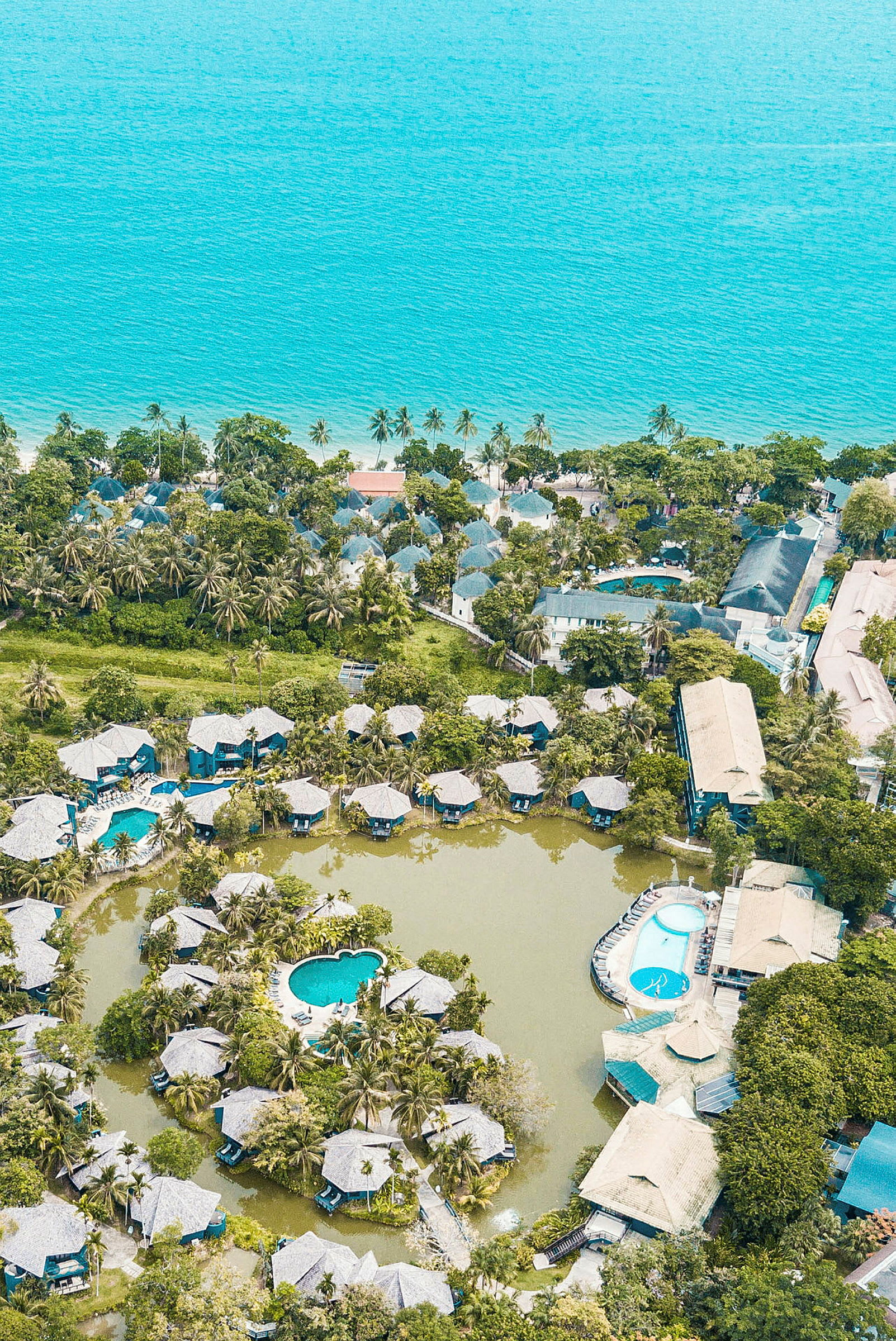 Flygfoto av en resort vid havet med bungalower kring pooler och grönska. (AI-genererad text)