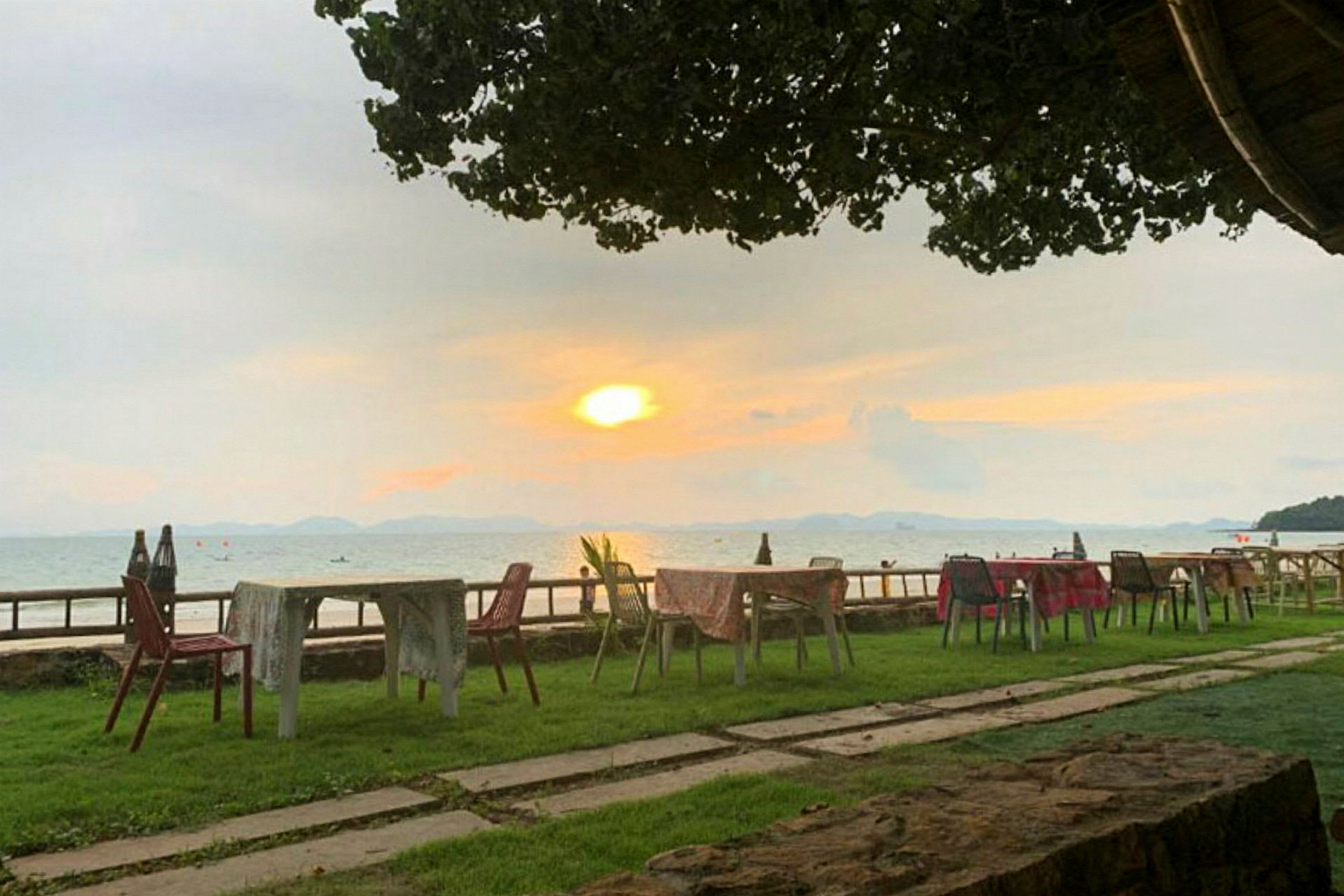 Uteservering med bord och stolar på gräsmatta vid havet med solnedgång i bakgrunden. (AI-genererad text)