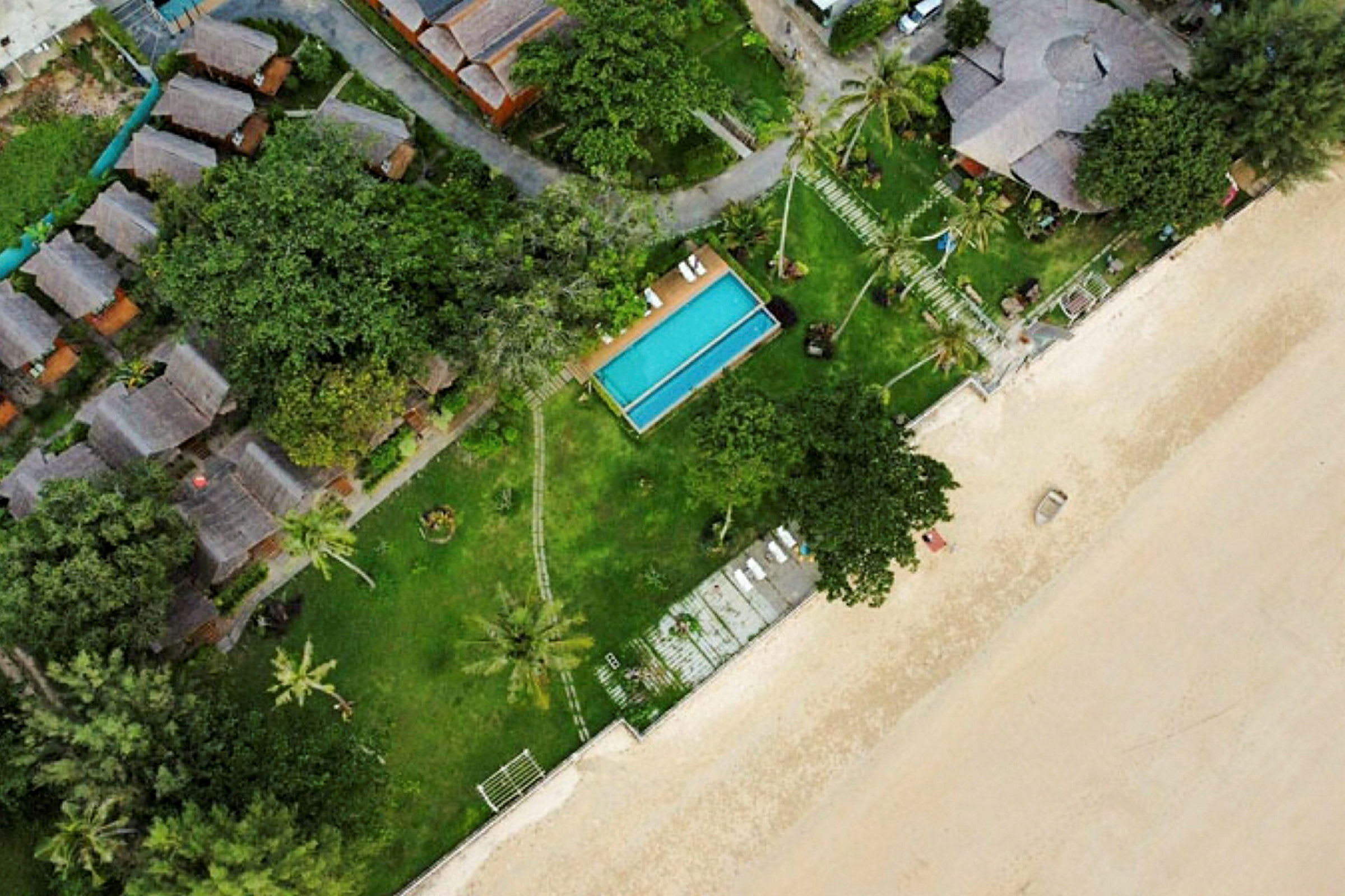 Flygfoto över en resort med flera byggnader och pool, omgiven av träd och belägen vid en sandstrand. (AI-genererad text)