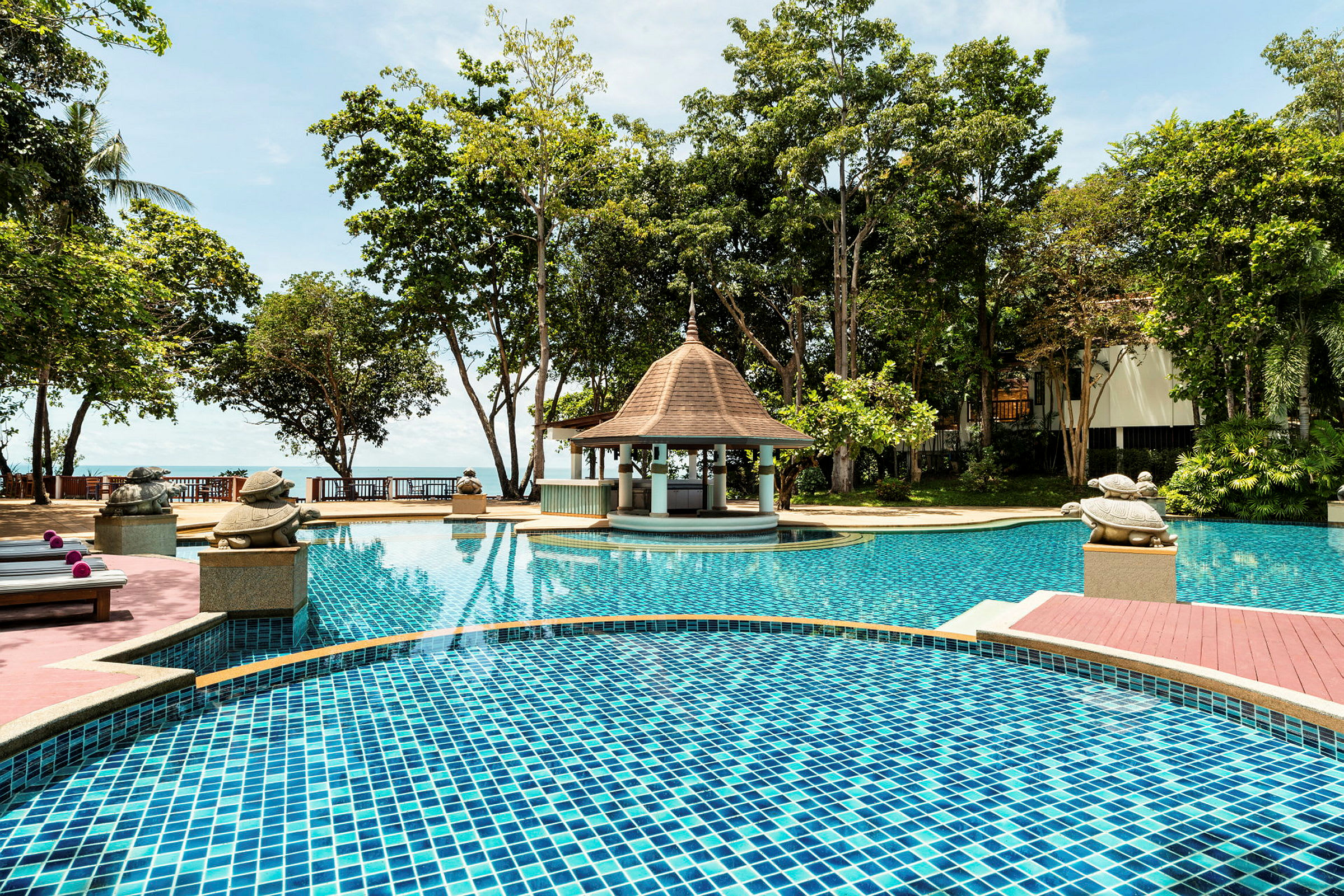 En pool vid ett hotellområde omgiven av träd, med ett litet paviljongliknande tak och dekorationer i form av sköldpaddor vid kanten. (AI-genererad text)