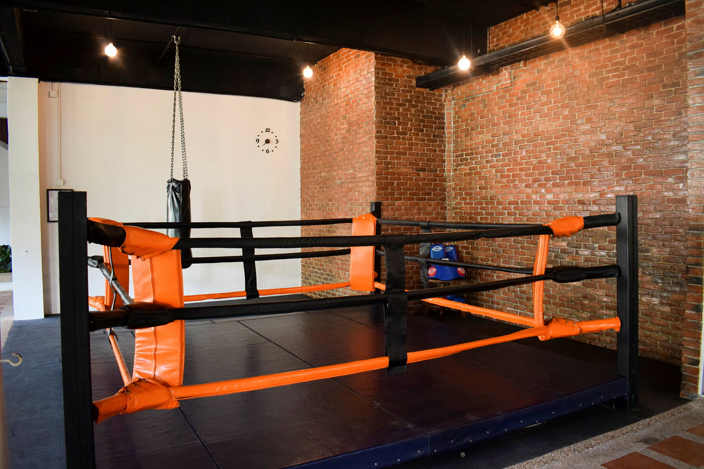 Ett rum med en boxningsring med orange och svarta skyddsstänger. Bakom ringen ses en hängande boxsäck och en vägg av tegelstenar. (AI-genererad text)