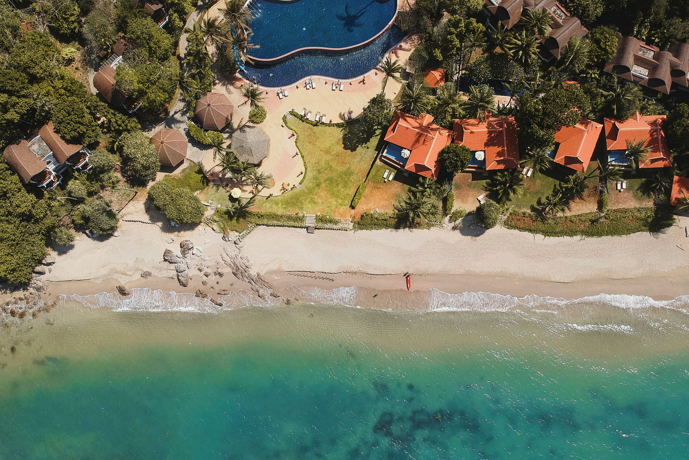 Flygfoto av en strand med turkost vatten, sand, stugor med röda tak och en swimmingpool. (AI-genererad text)