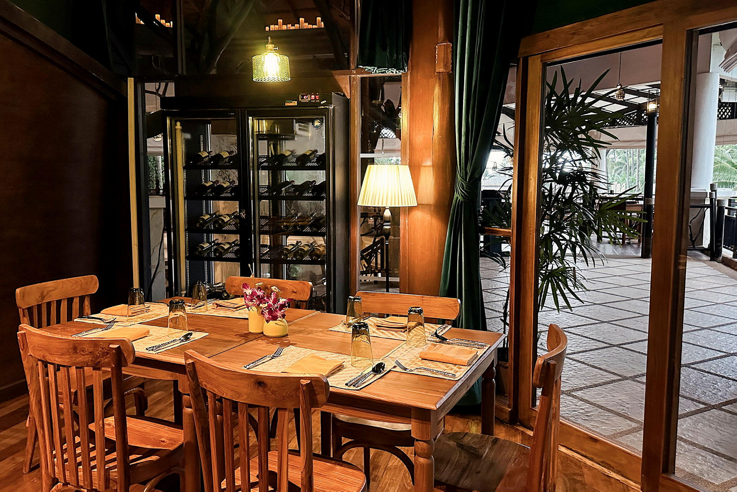 Ett träbord med trästolar är dukat för fyra personer i en restaurangmiljö. Vinhyllor och gröna draperier är synliga i bakgrunden. (AI-genererad text)