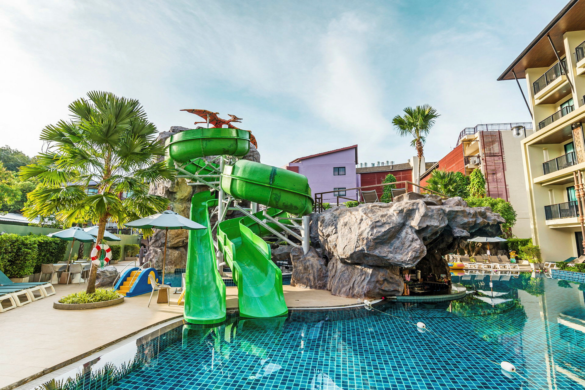 Grön vattenrutschkana vid en pool, omgiven av solstolar och byggnader. (AI-genererad text)