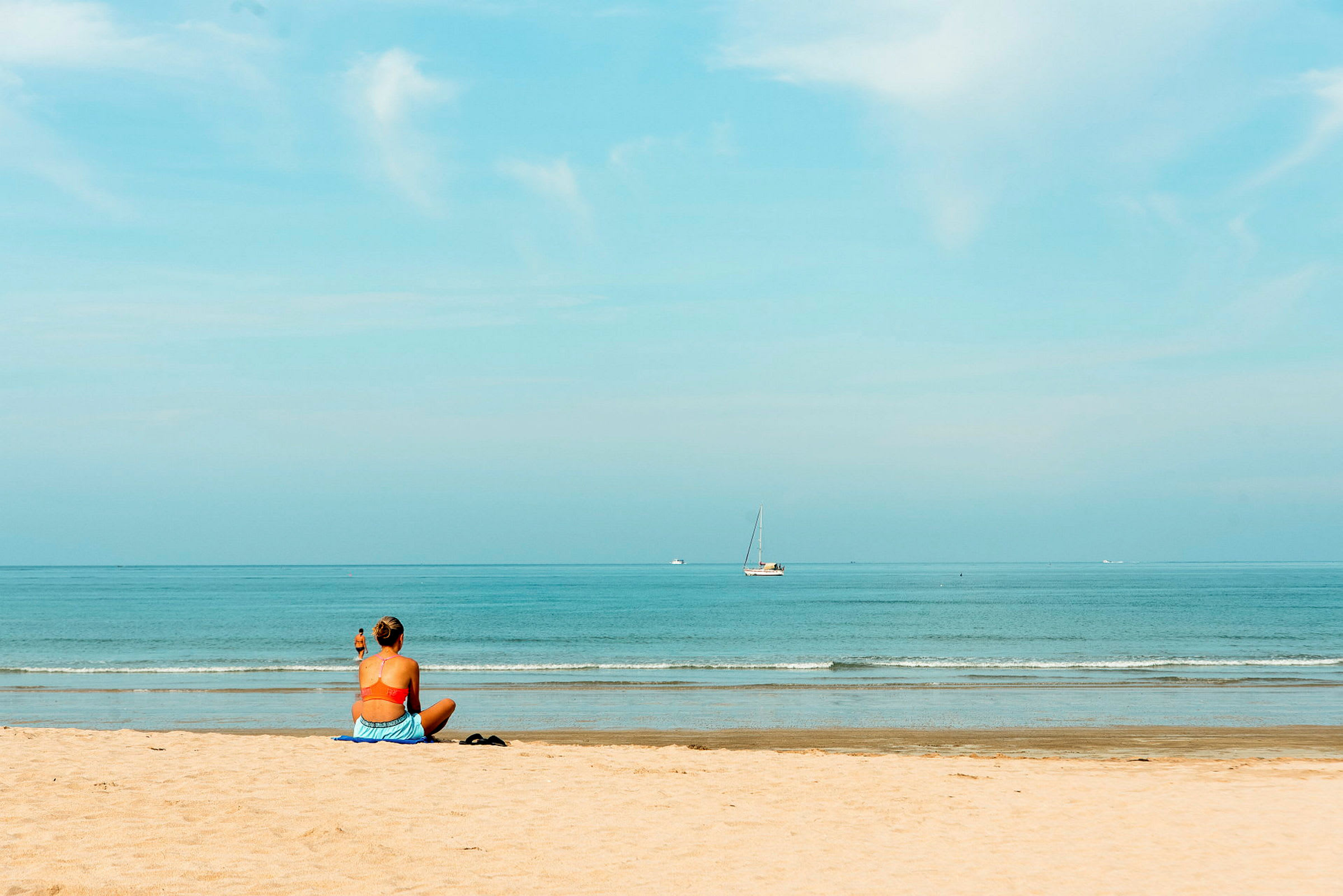 En person sitter på en strand och ser ut över havet, där en segelbåt syns i fjärran. (AI-genererad text)