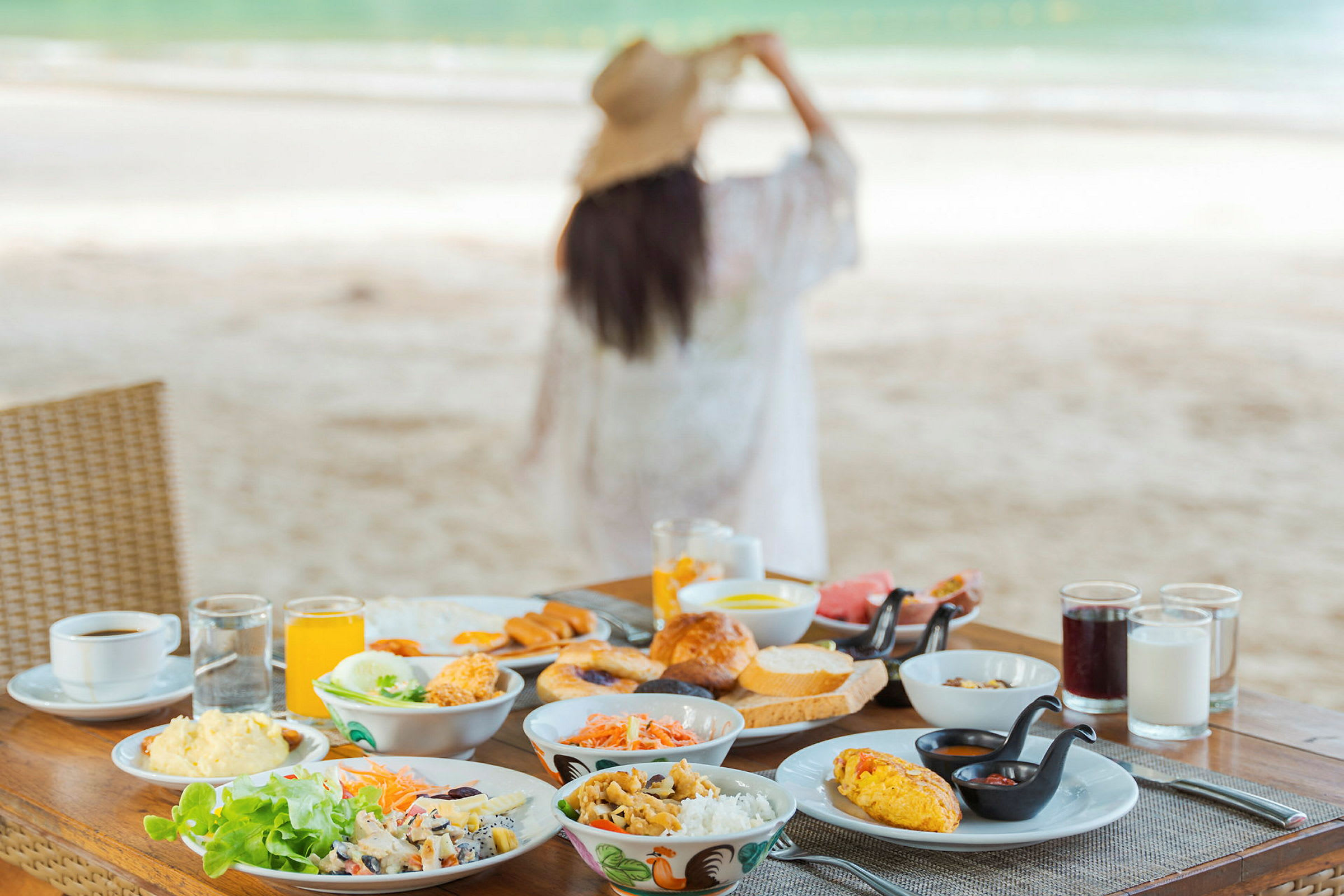 Ett frukostbord med diverse maträtter och drycker står på en strand. I bakgrunden syns en person som tittar ut över havet. (AI-genererad text)