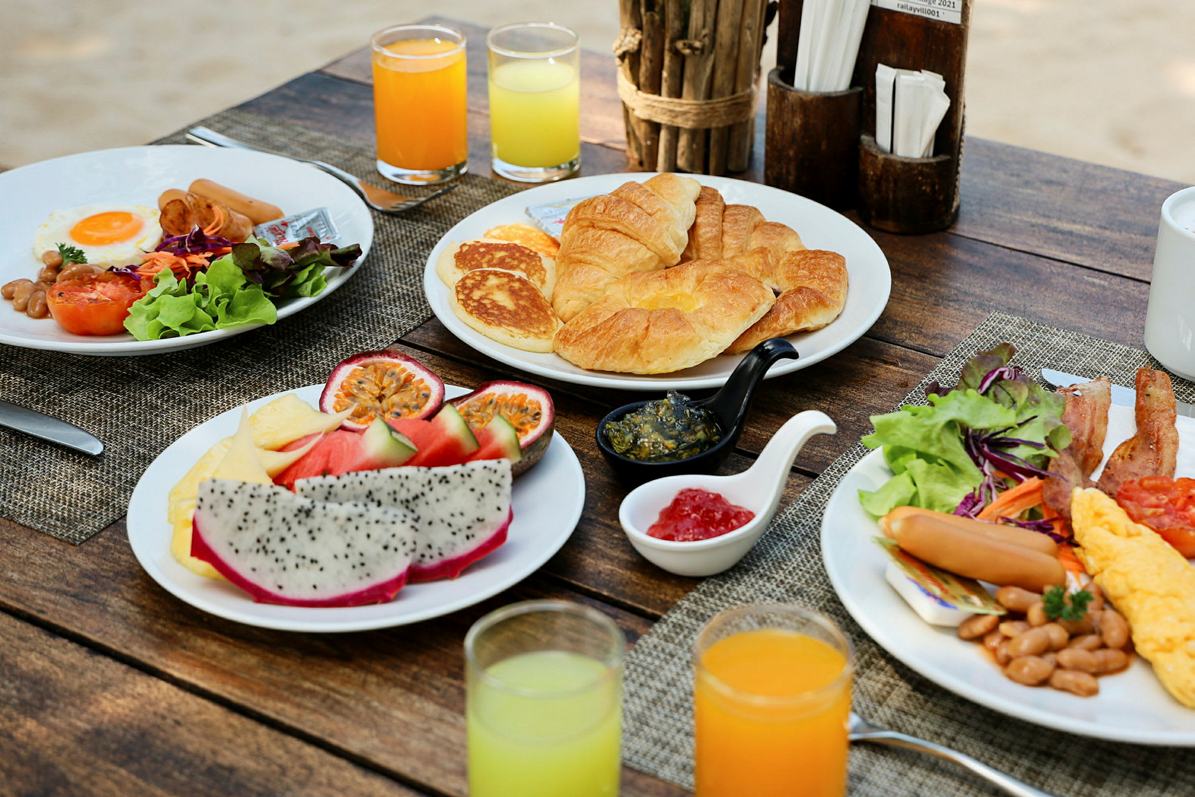 En frukostuppsättning med olika rätter serverad på ett rustikt träbord, inklusive croissanter, pannkakor, frukt, ägg och sallad, med juice i glas. (AI-genererad text)