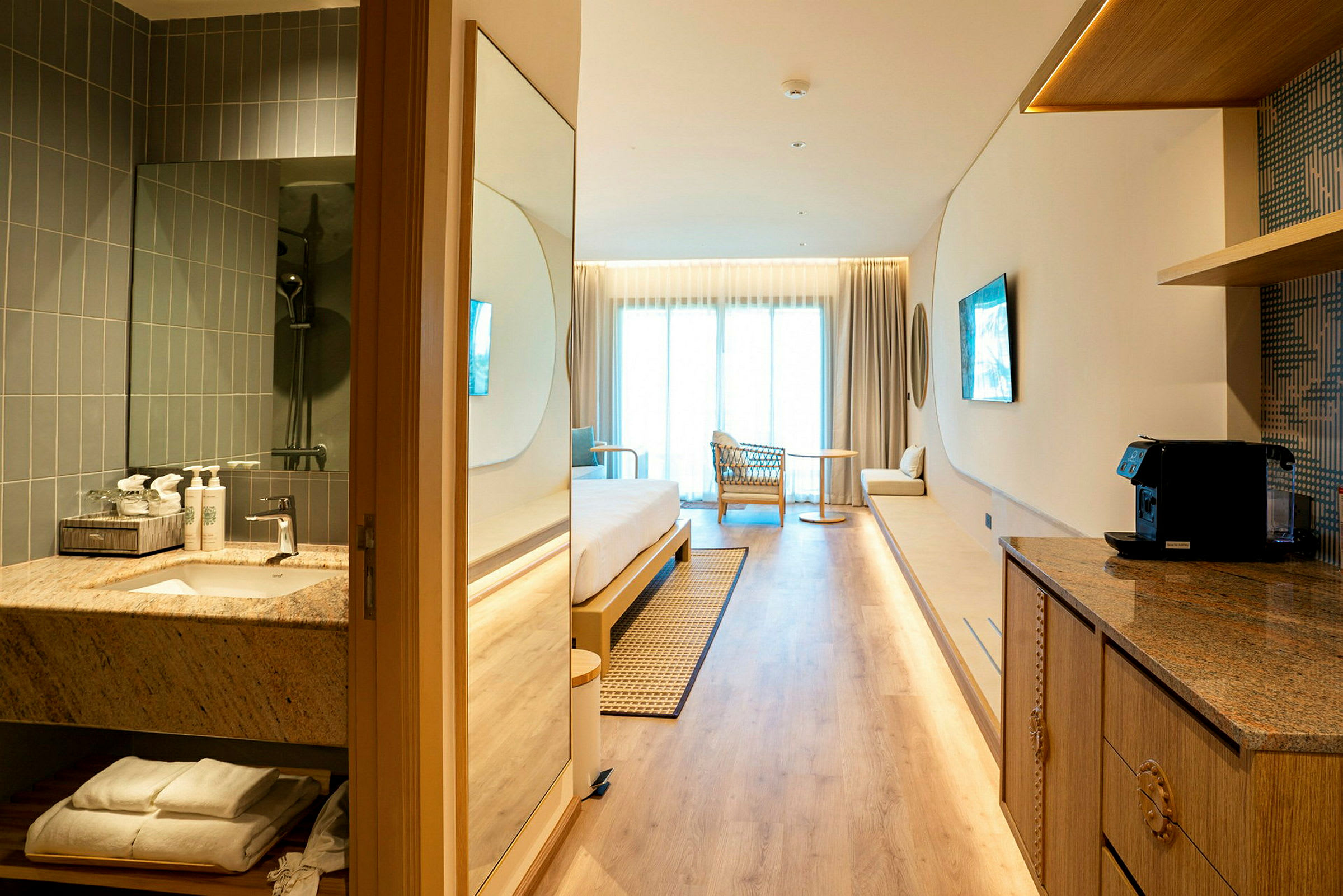 Ett modernt hotellrum med ljus inredning, inklusive säng, fåtölj, litet bord och balkong. Ett badrum och en köksdel syns i förgrunden. (AI-genererad text)