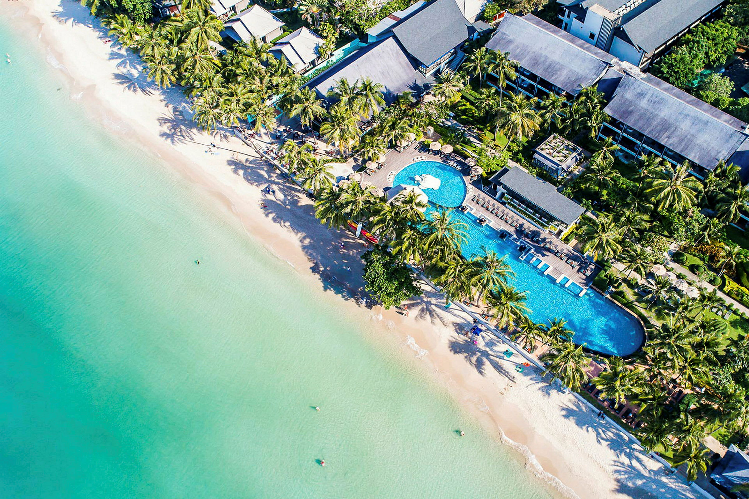 Flygbild av en resort vid stranden, med swimmingpool, palmer och byggnader. (AI-genererad text)