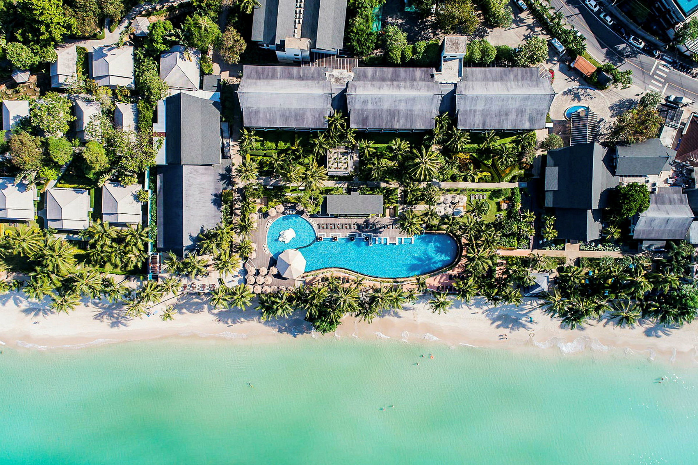 Flygbild av ett semesterresort med pool, omgiven av tropiska träd och nära en strand med klart vatten. (AI-genererad text)