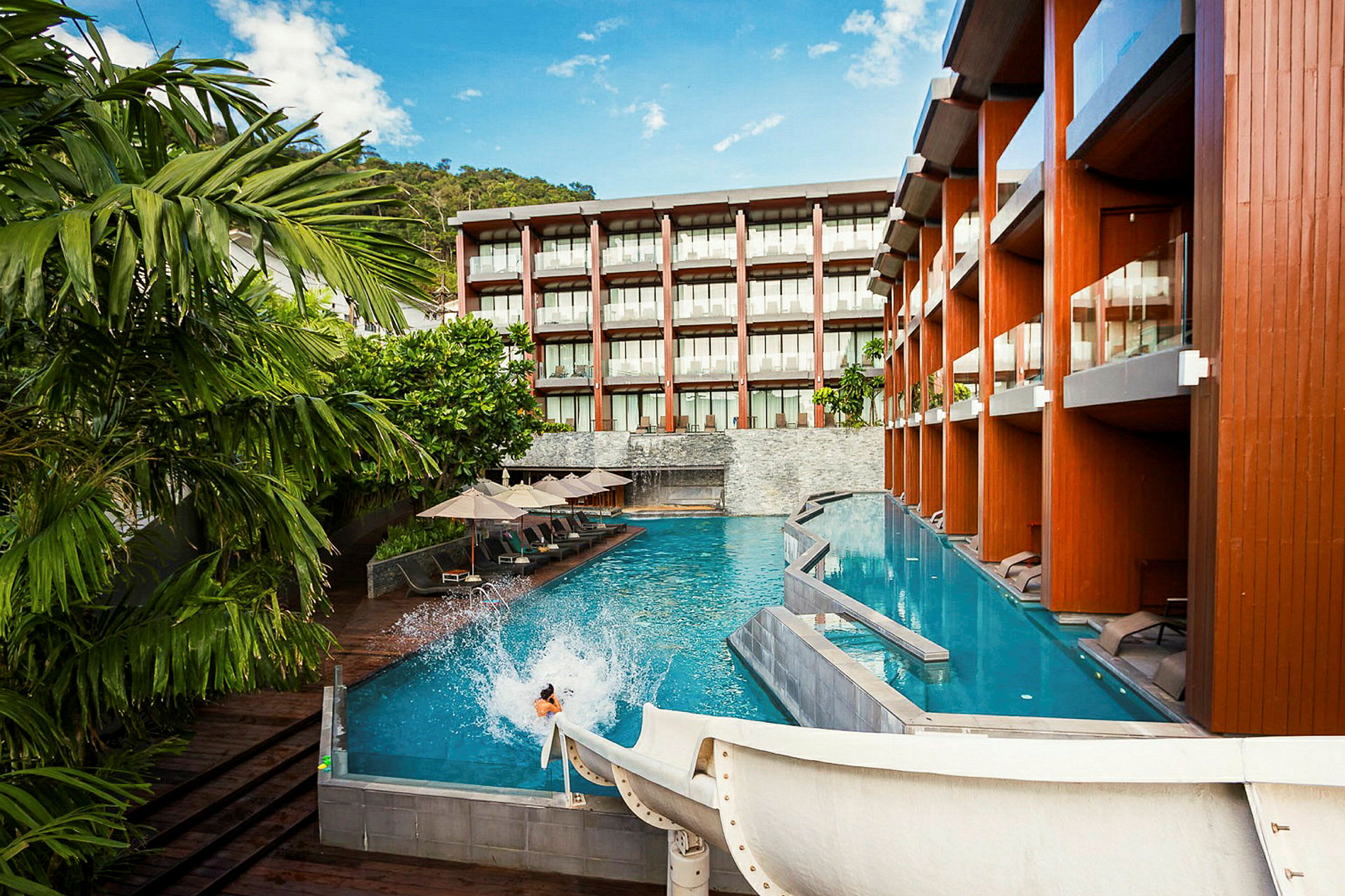 En pool vid ett modernt hotell med solstolar och parasoller samt en vattenrutschkana som leder ner i vattnet. (AI-genererad text)