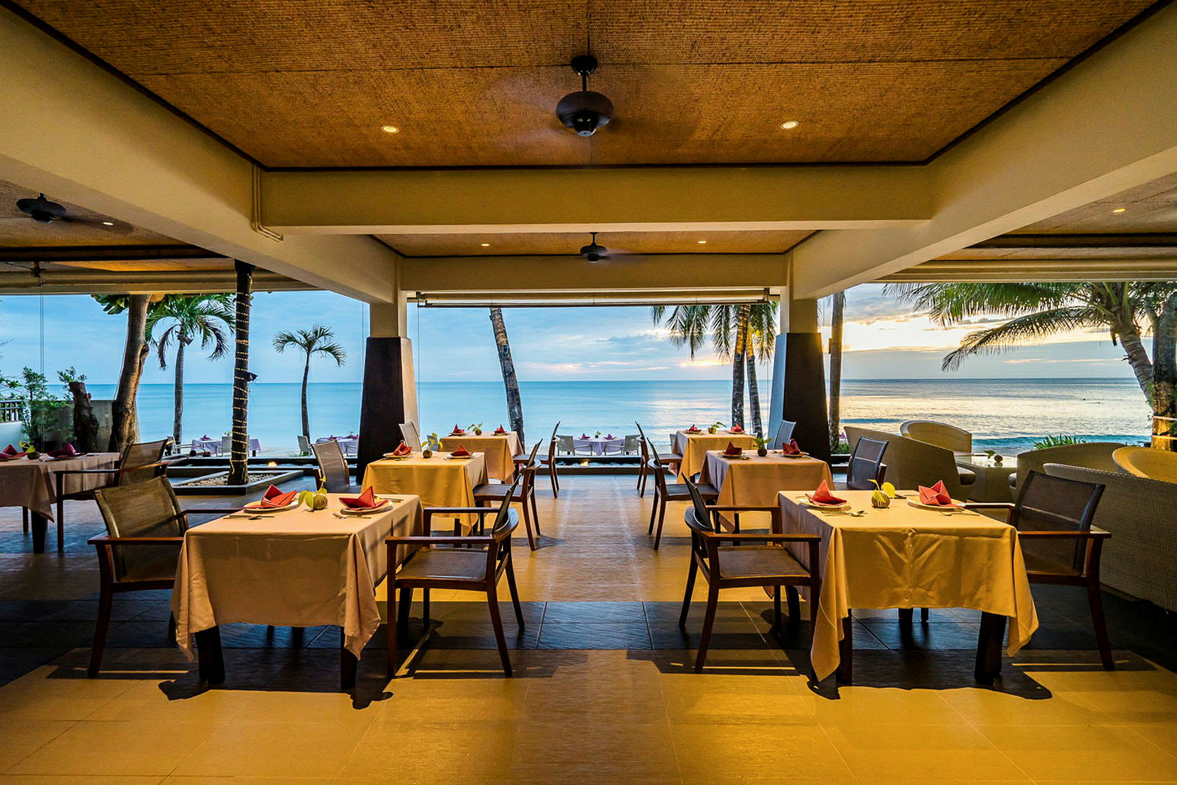 En restaurang vid havet med vita dukar på borden och en utsikt över palmer och stranden. (AI-genererad text)