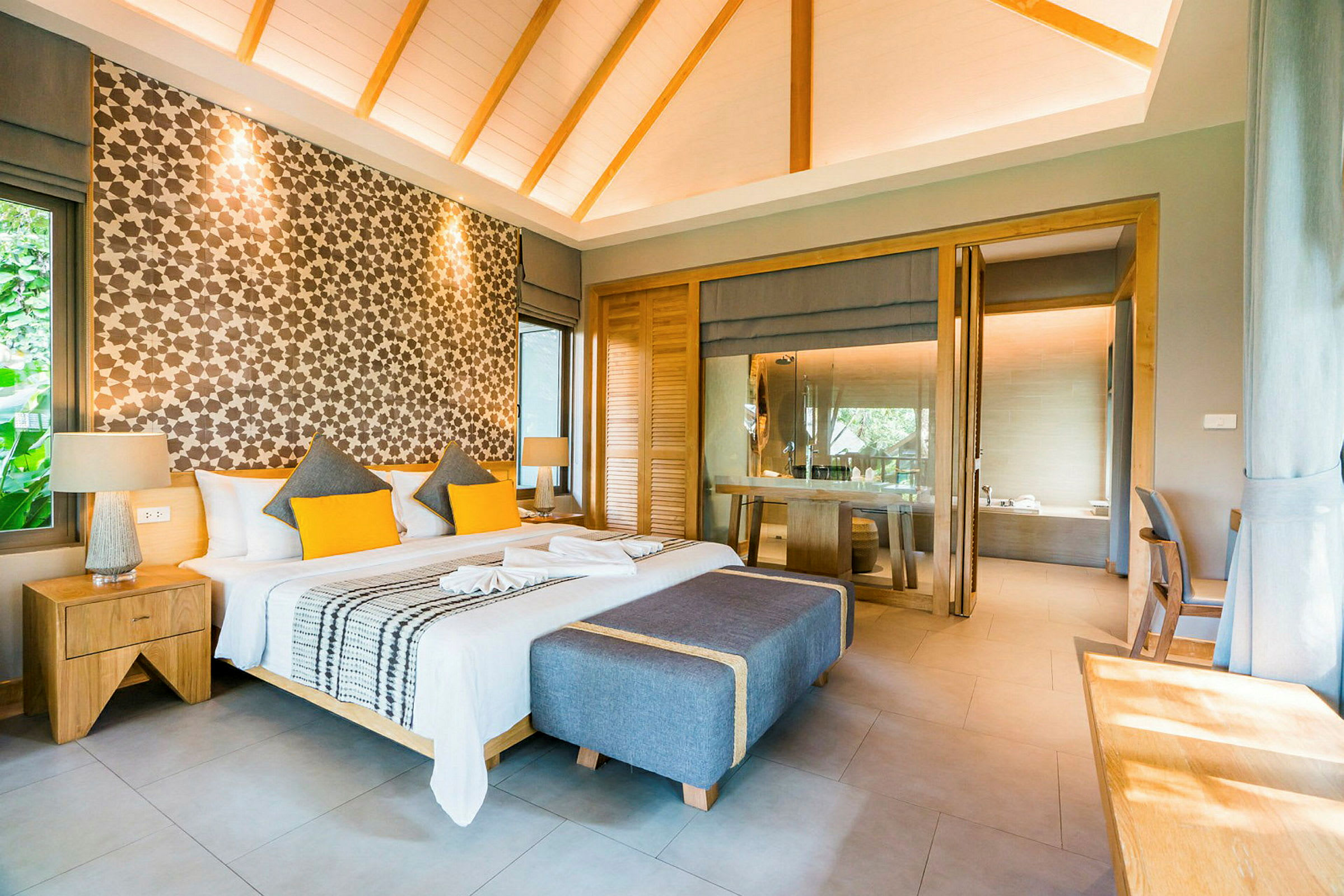 Ett modernt och stilfullt hotellrum med en stor dubbelsäng, dekorativa kuddar och mönstrad väggbakgrund. Roommet har också fönster som släpper in naturligt ljus och är inrett med trädetaljer. (AI-genererad text)